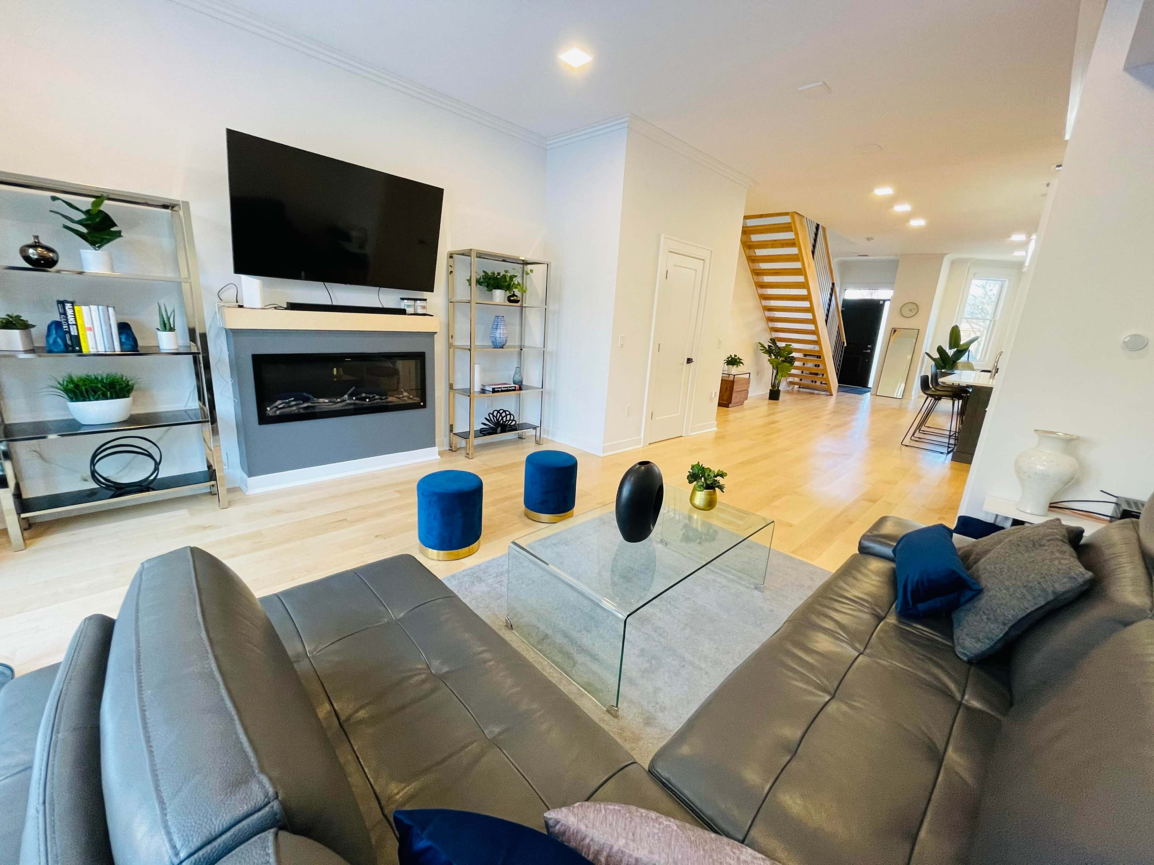 A modern living room with a large flat-screen TV on the wall, a glass coffee table, gray sofas, and a staircase in the background.