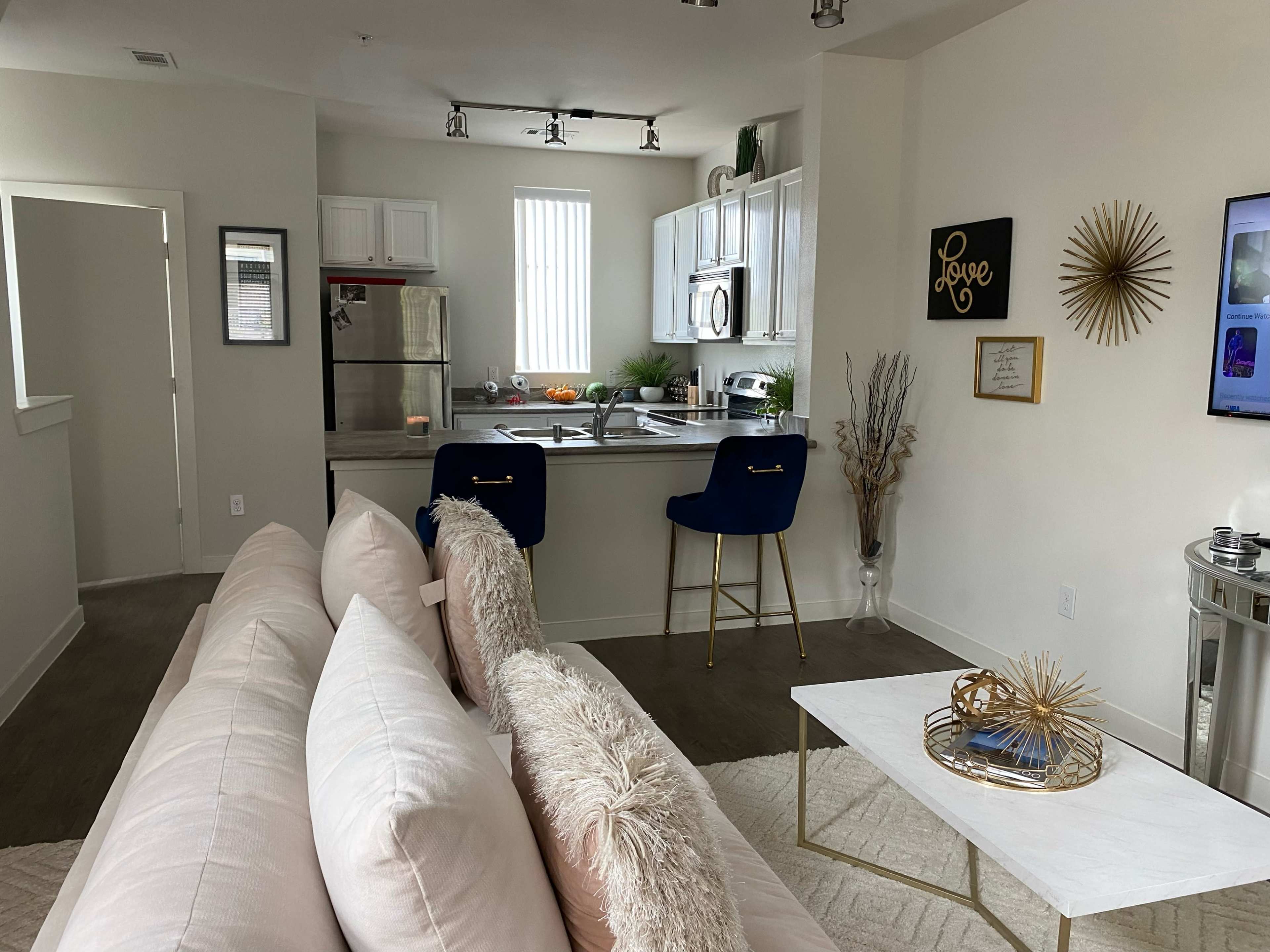 The image shows a modern living space featuring a kitchen area with white cabinets, blue bar stools, and a light-colored sofa in the foreground.