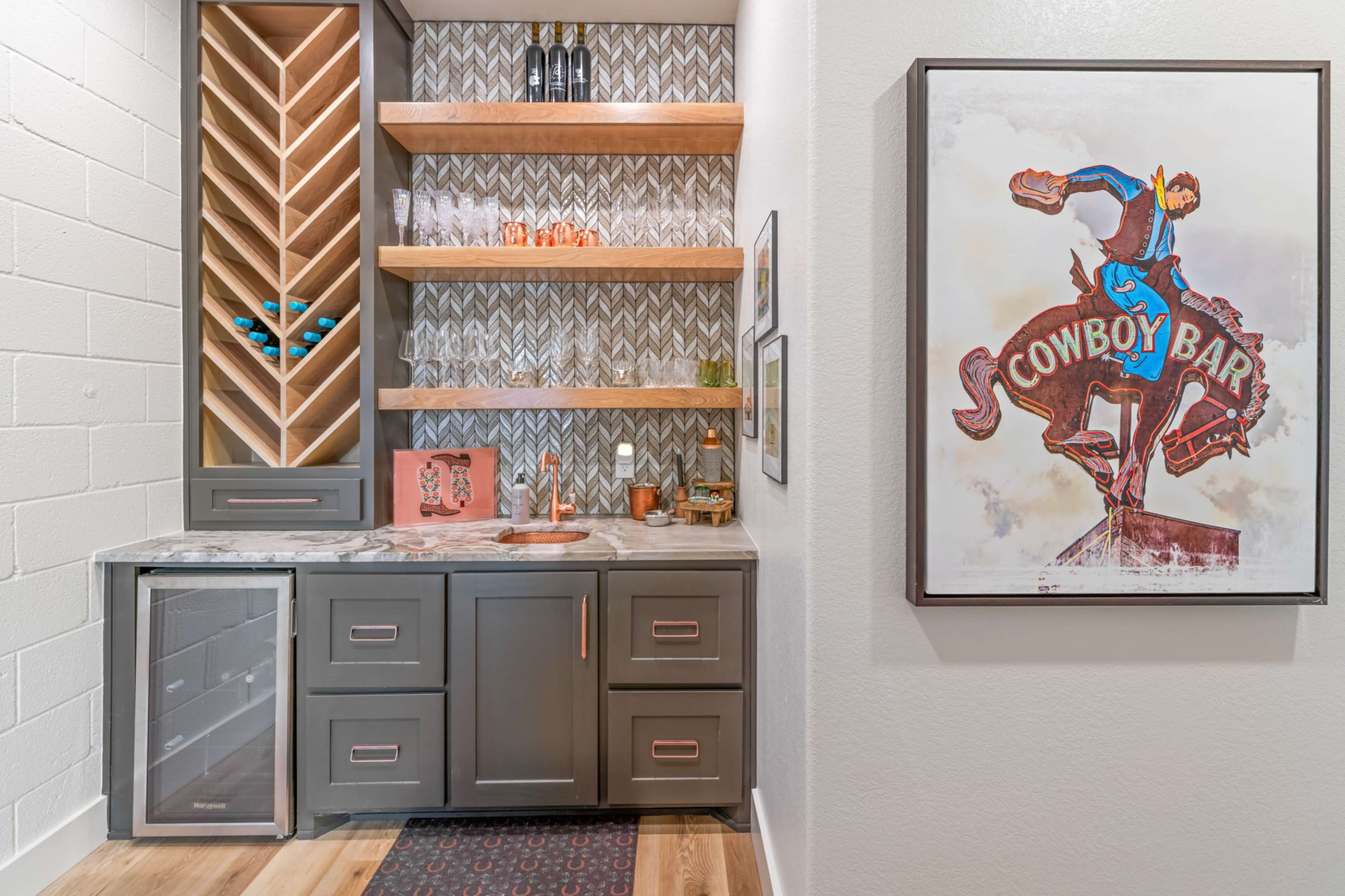 The image shows a modern bar area featuring a wine rack, open shelves with glassware, a countertop with a sink, and a framed cowboy-themed artwork on the wall.