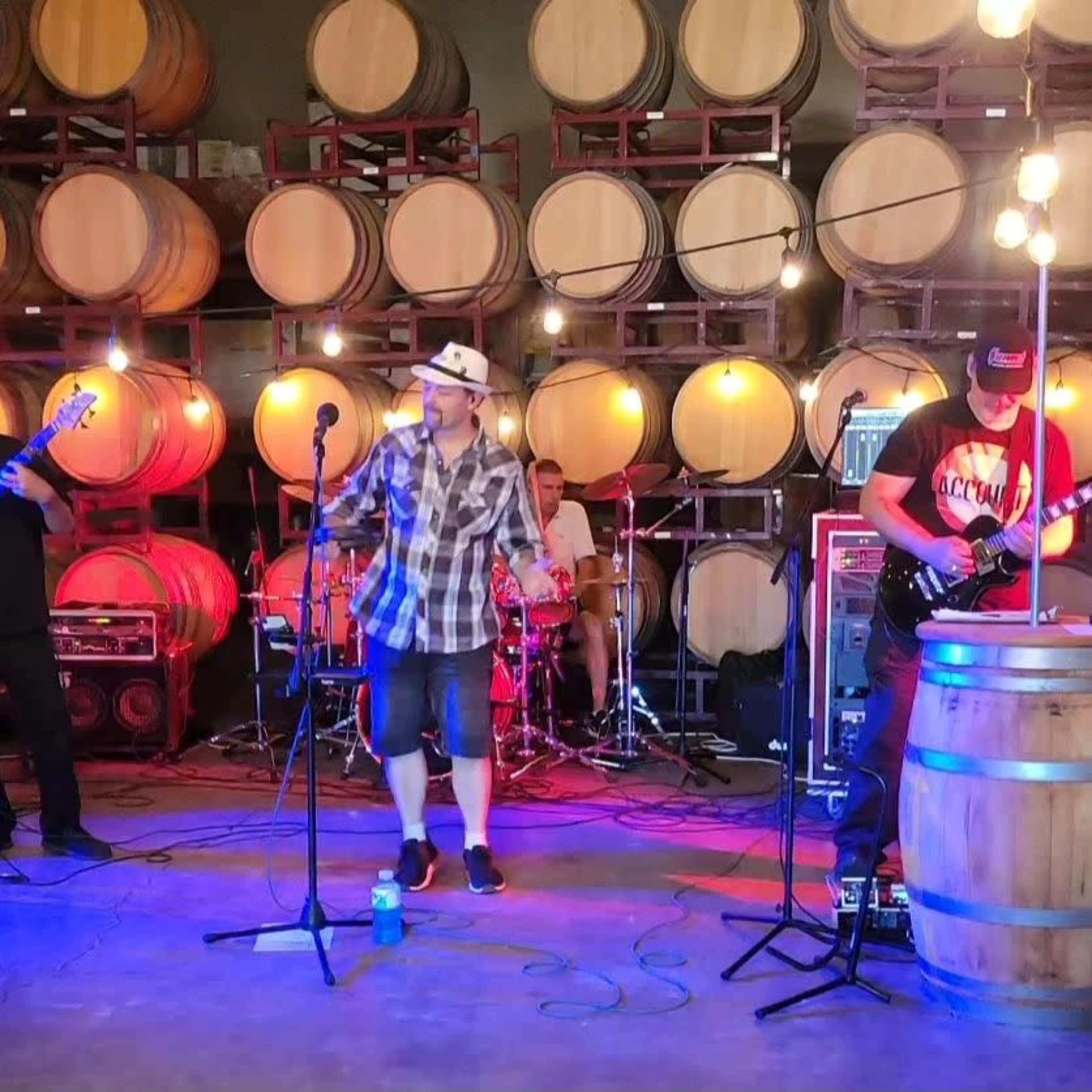 A band performs on stage in a winery, surrounded by large wine barrels and illuminated by overhead lights.