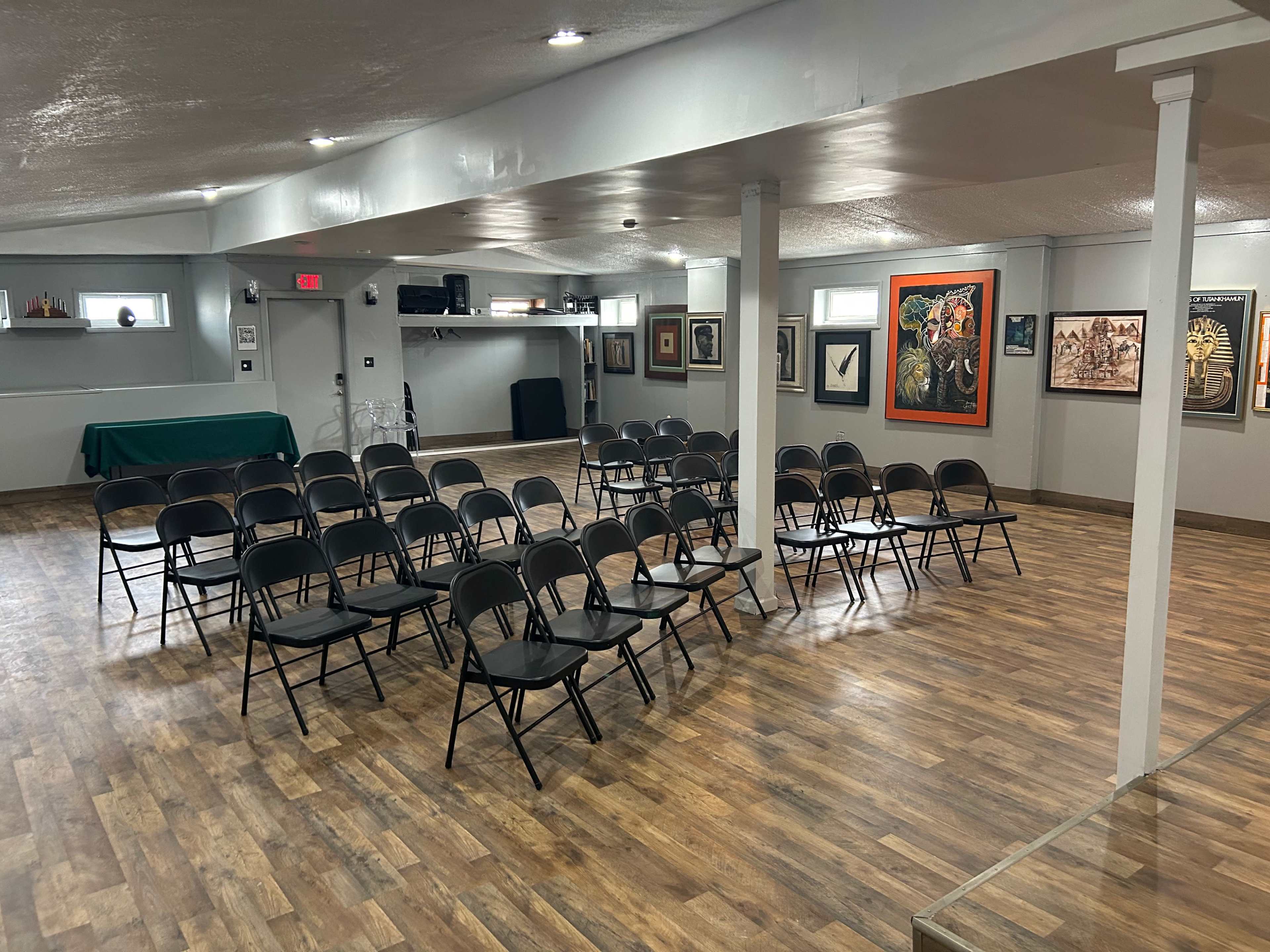 A spacious room set up with rows of black chairs facing a stage area, accompanied by various framed artworks on the walls.