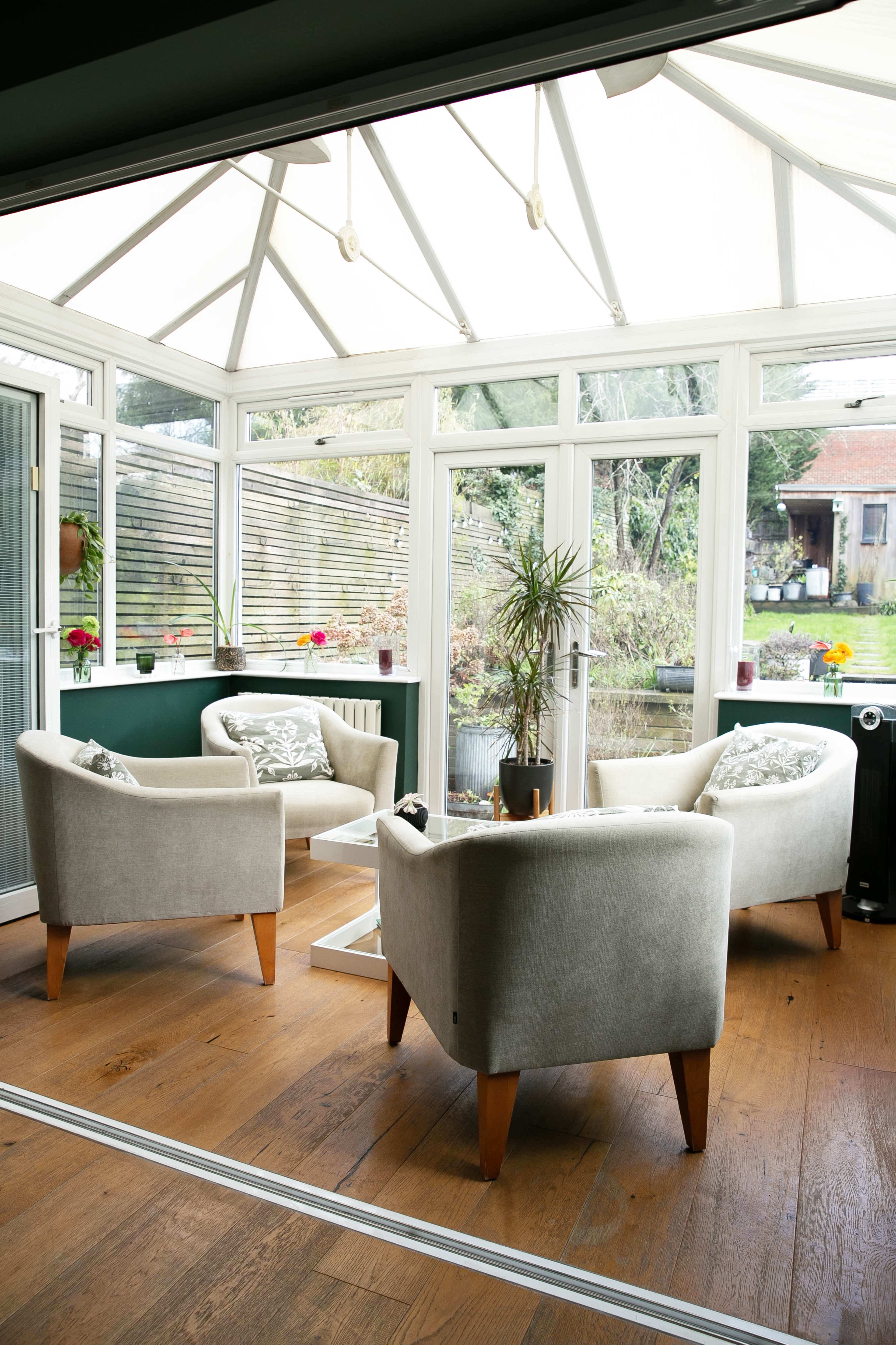 The image shows a sunlit conservatory with two beige armchairs and a small table, surrounded by greenery and flowers in the windows.