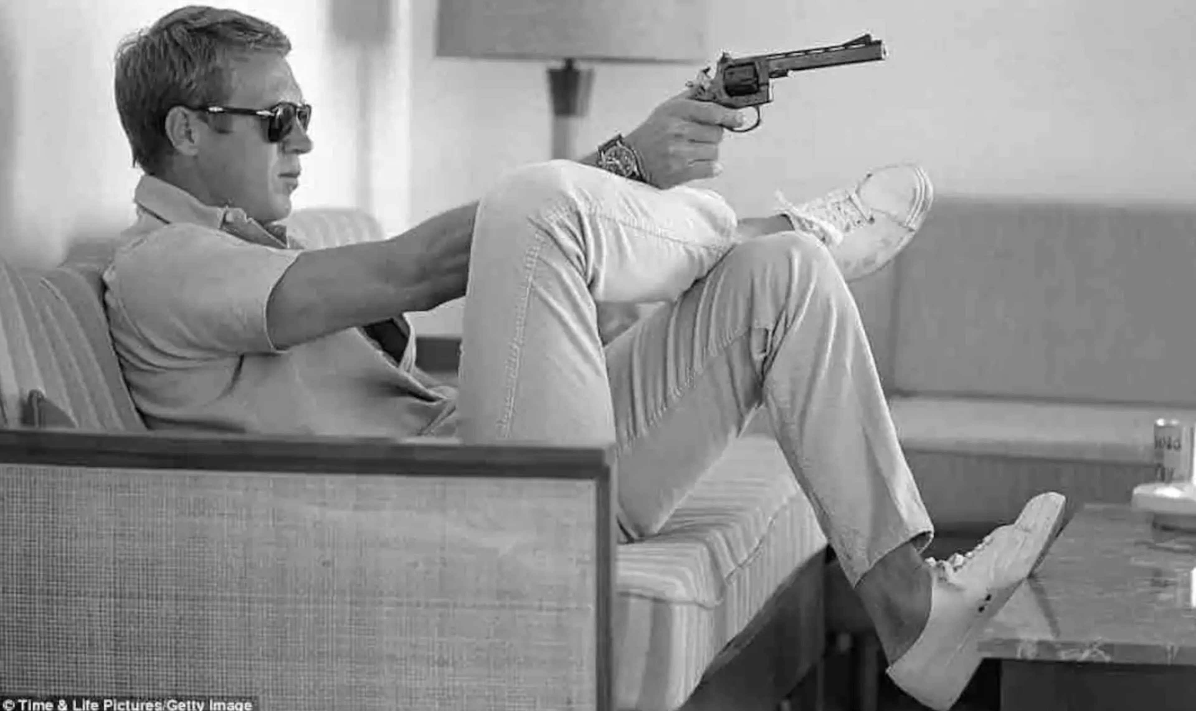 A man sits on a couch with his legs crossed, holding a handgun and wearing sunglasses.