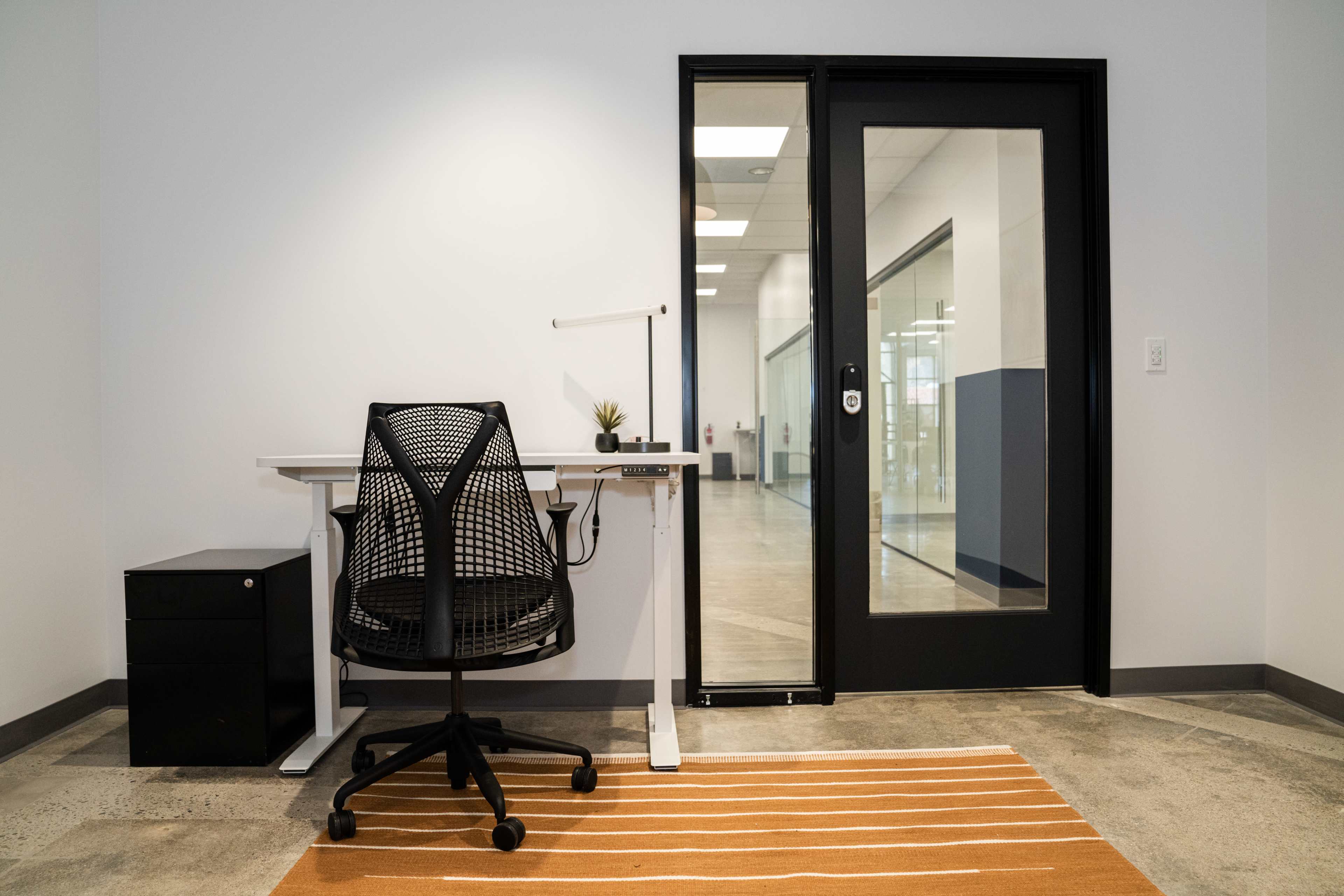 A minimalist office space featuring a desk with a chair, a small filing cabinet, and a doorway leading to another room.