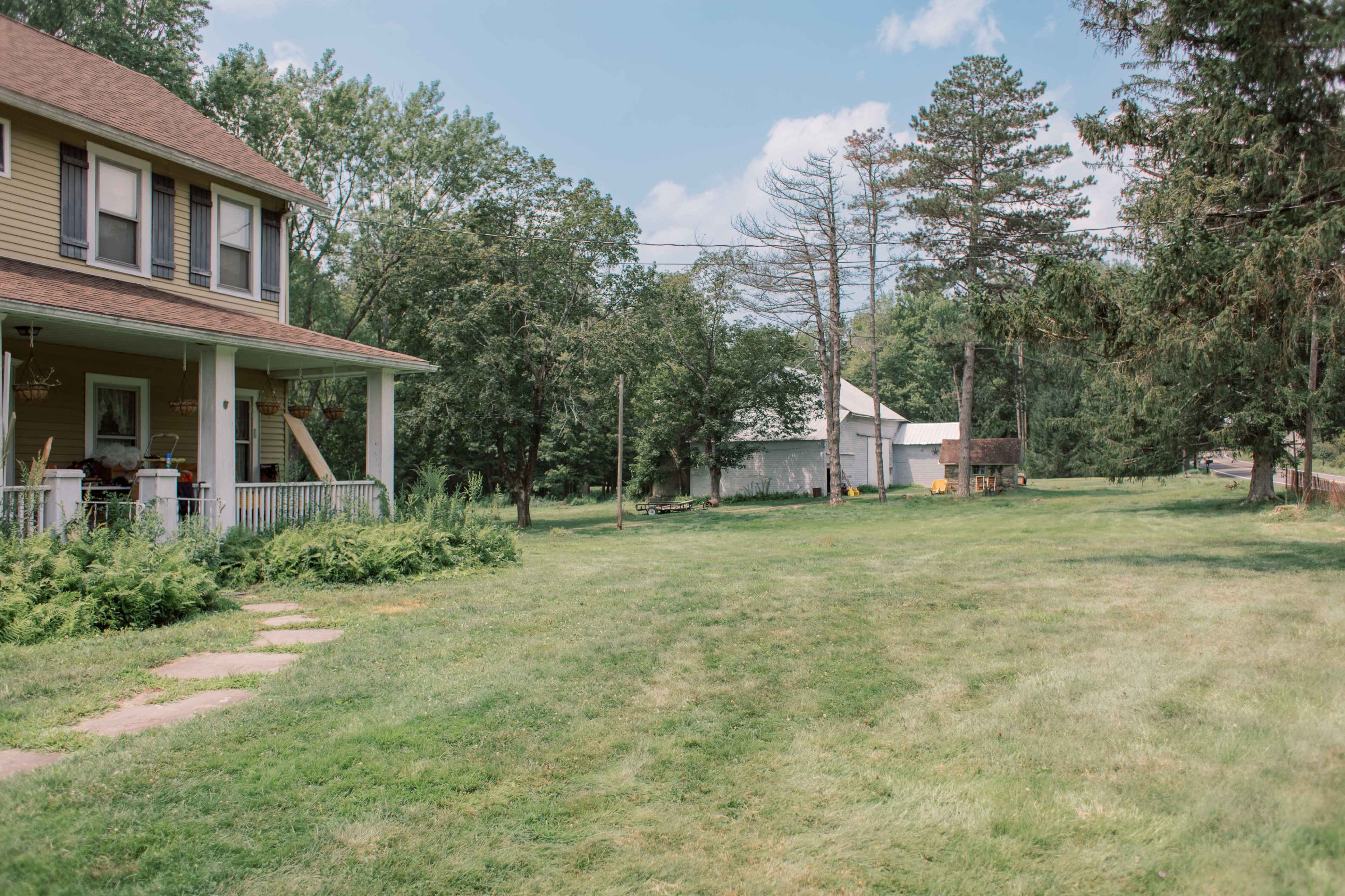 A yellow two-story house with a porch sits on a grassy lawn, surrounded by trees and a white shed in the background.