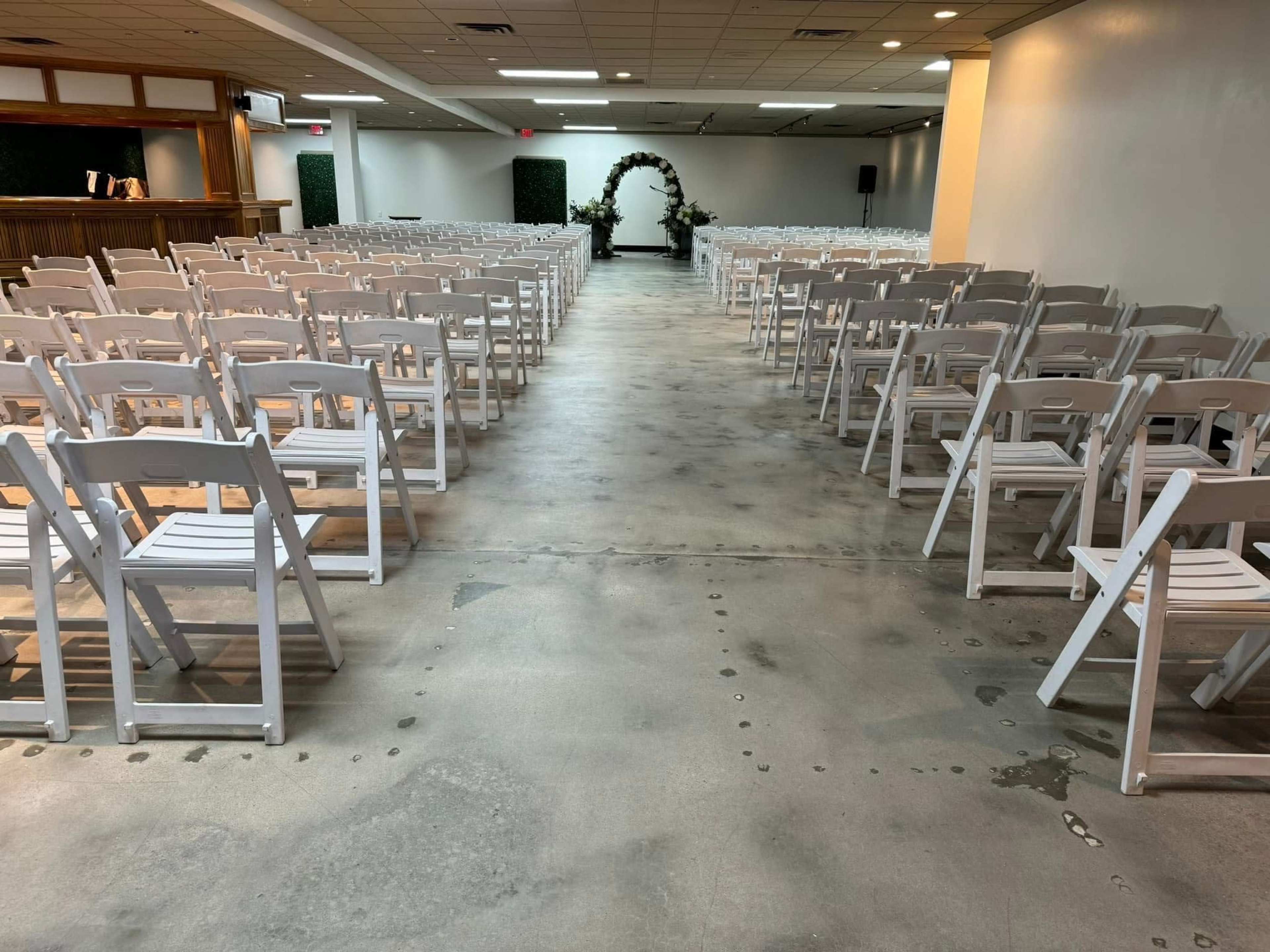 The image shows a spacious indoor venue set up with rows of white folding chairs facing a decorated archway at the front.