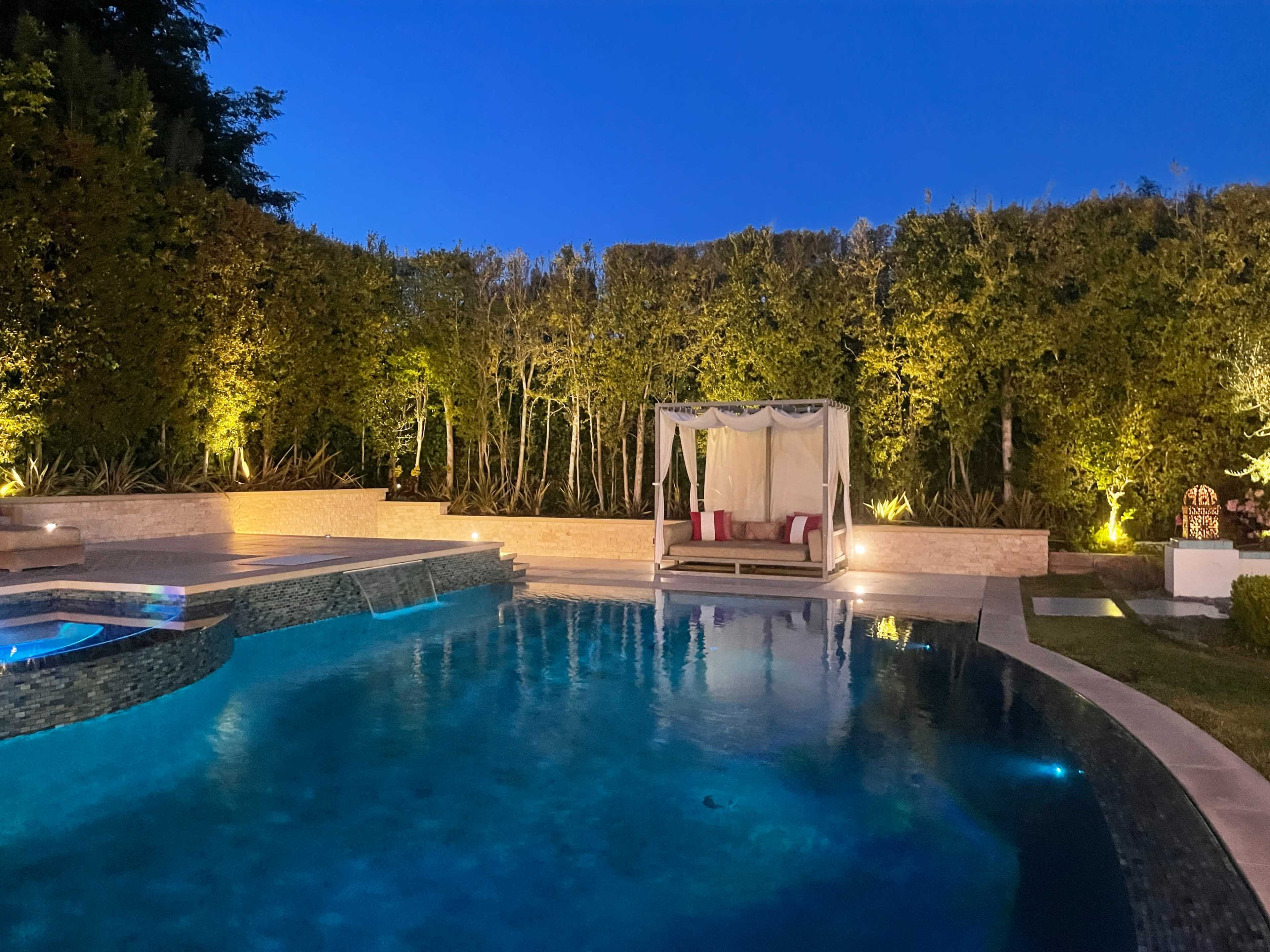 A well-lit outdoor pool area with lounge seating under a canopy, surrounded by greenery at dusk.
