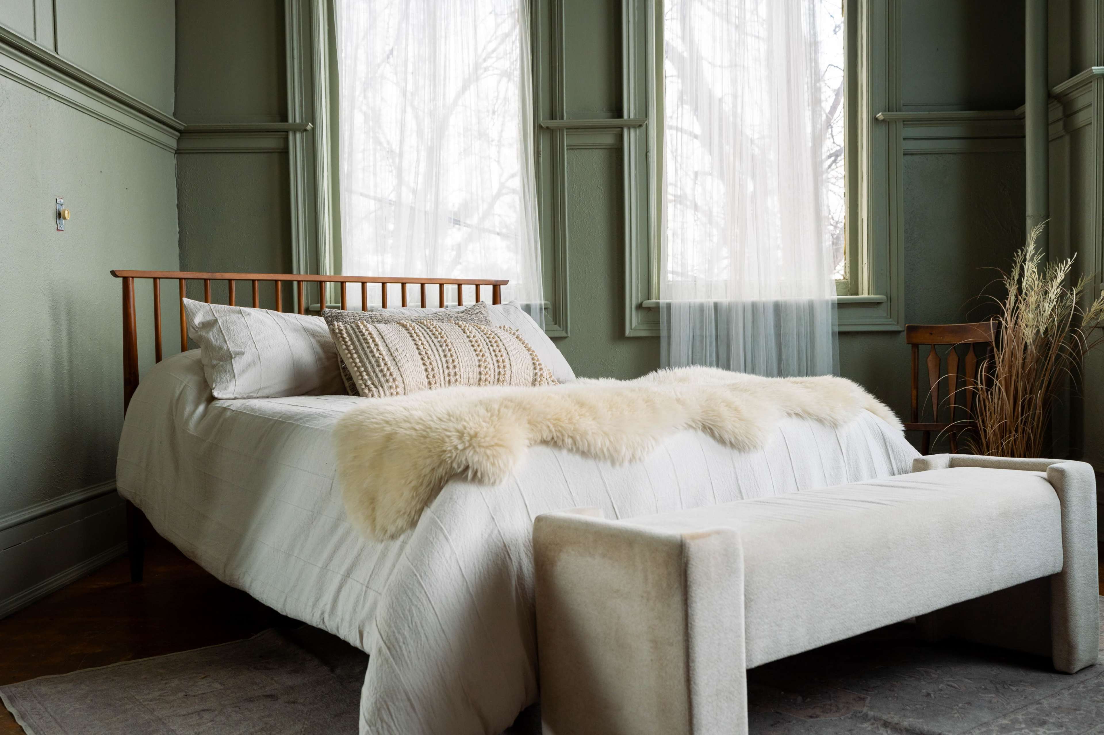 A neatly made bed with a white quilt and decorative pillows is positioned in a softly lit bedroom featuring large windows and a beige bench at the foot.