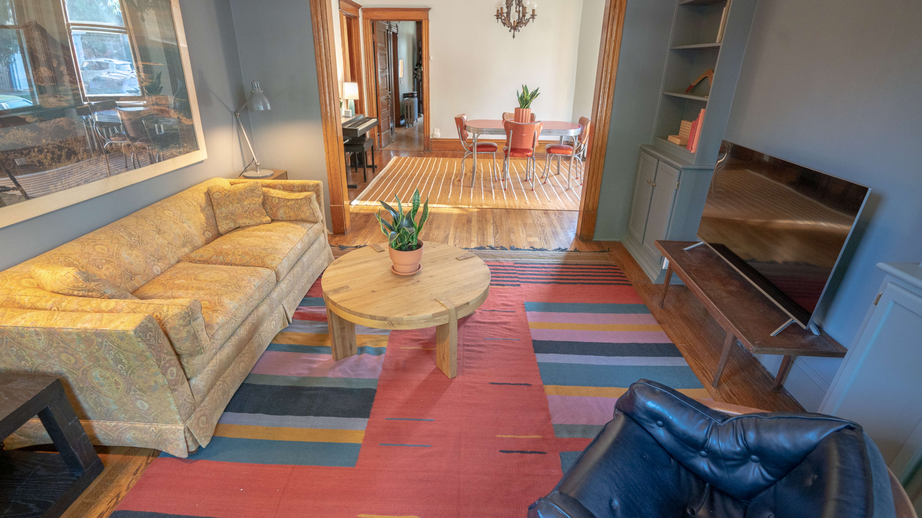 A living room with a yellow floral sofa, a round wooden coffee table, a black armchair, and a television on a wooden stand, with an adjacent dining area visible in the background.
