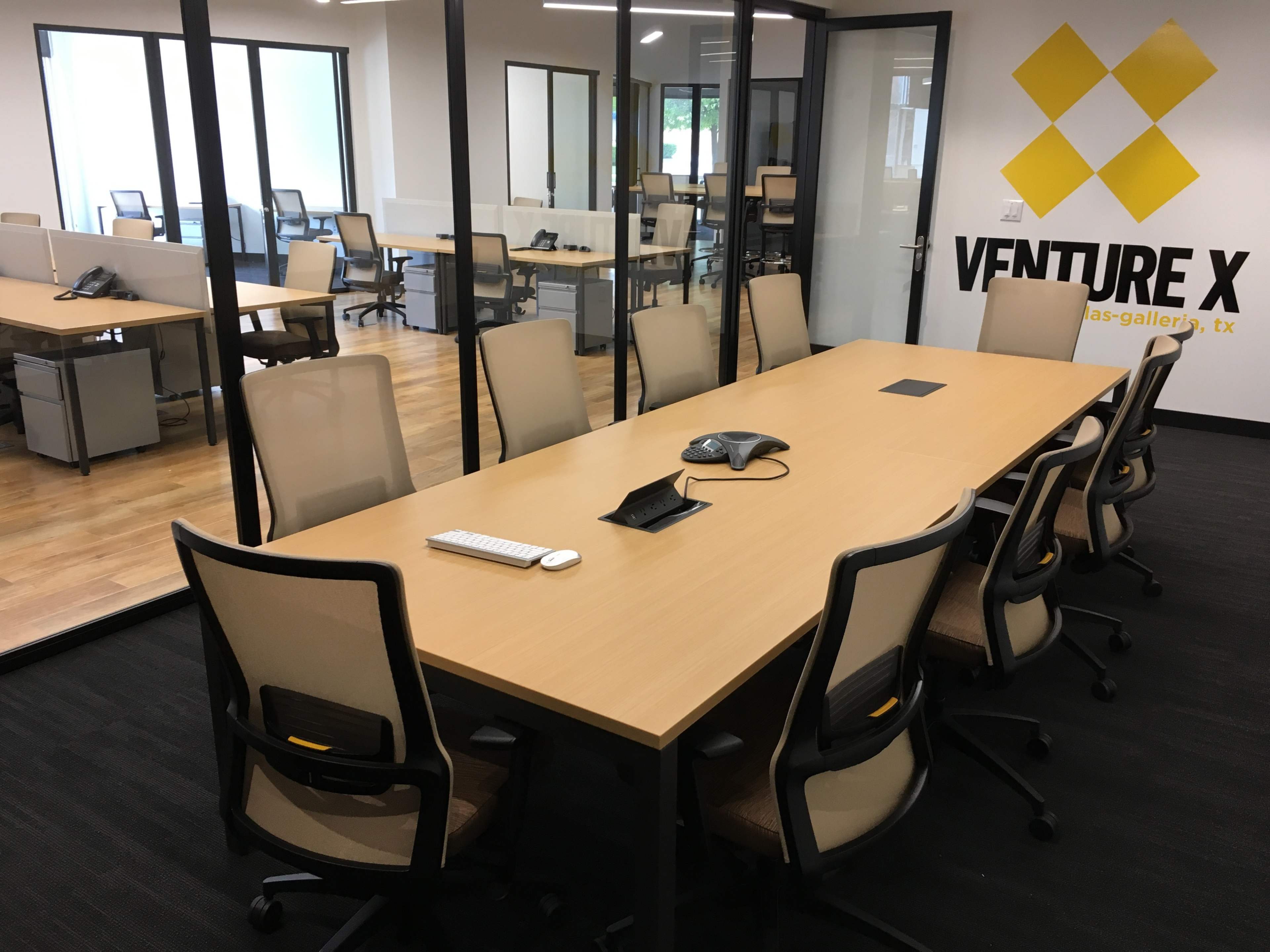 A spacious conference room features a long wooden table surrounded by ergonomic chairs, with a phone and keyboard on the table, and glass partitions revealing additional workspaces.