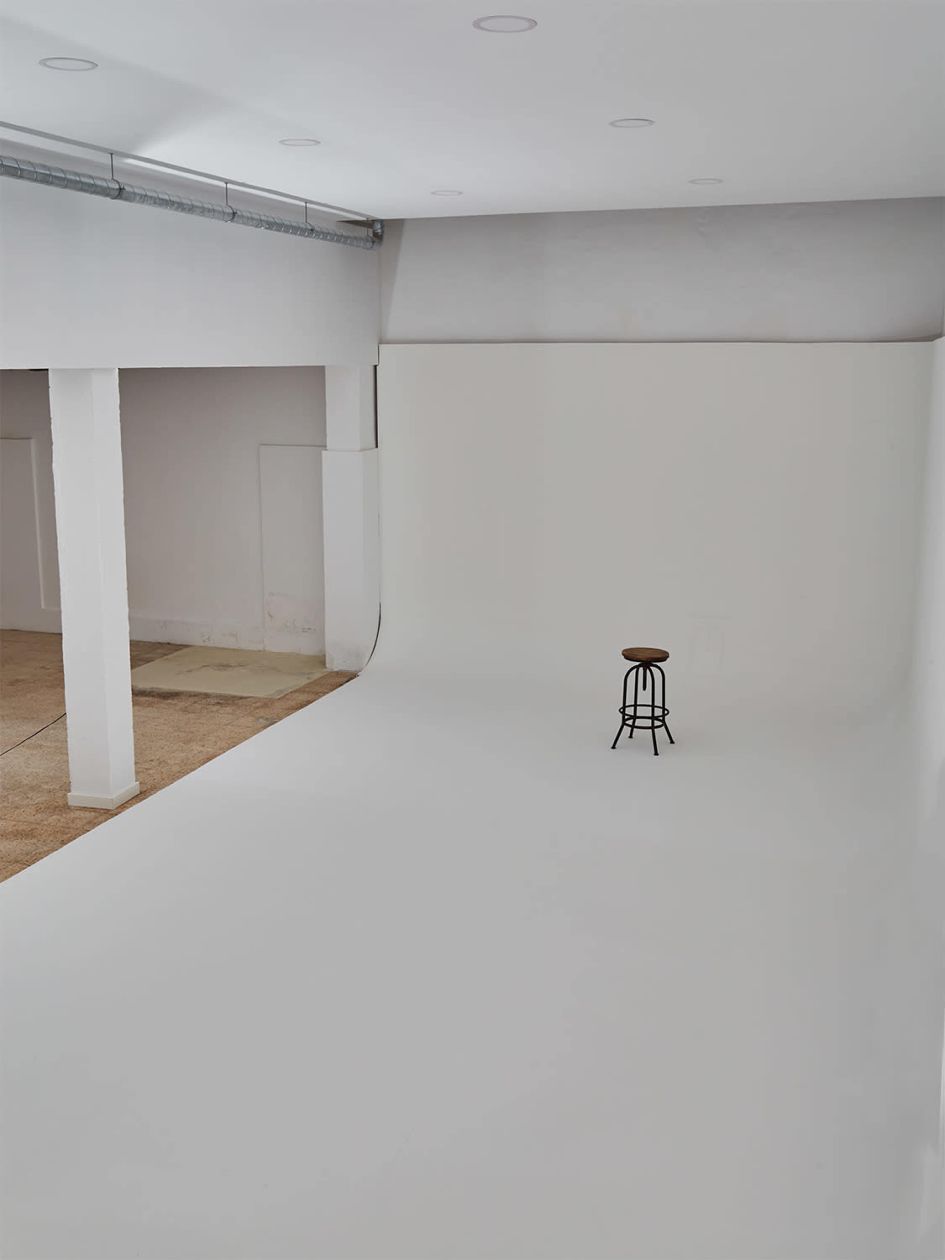 A minimalistic photography studio features a smooth white backdrop with a black stool positioned in the center.