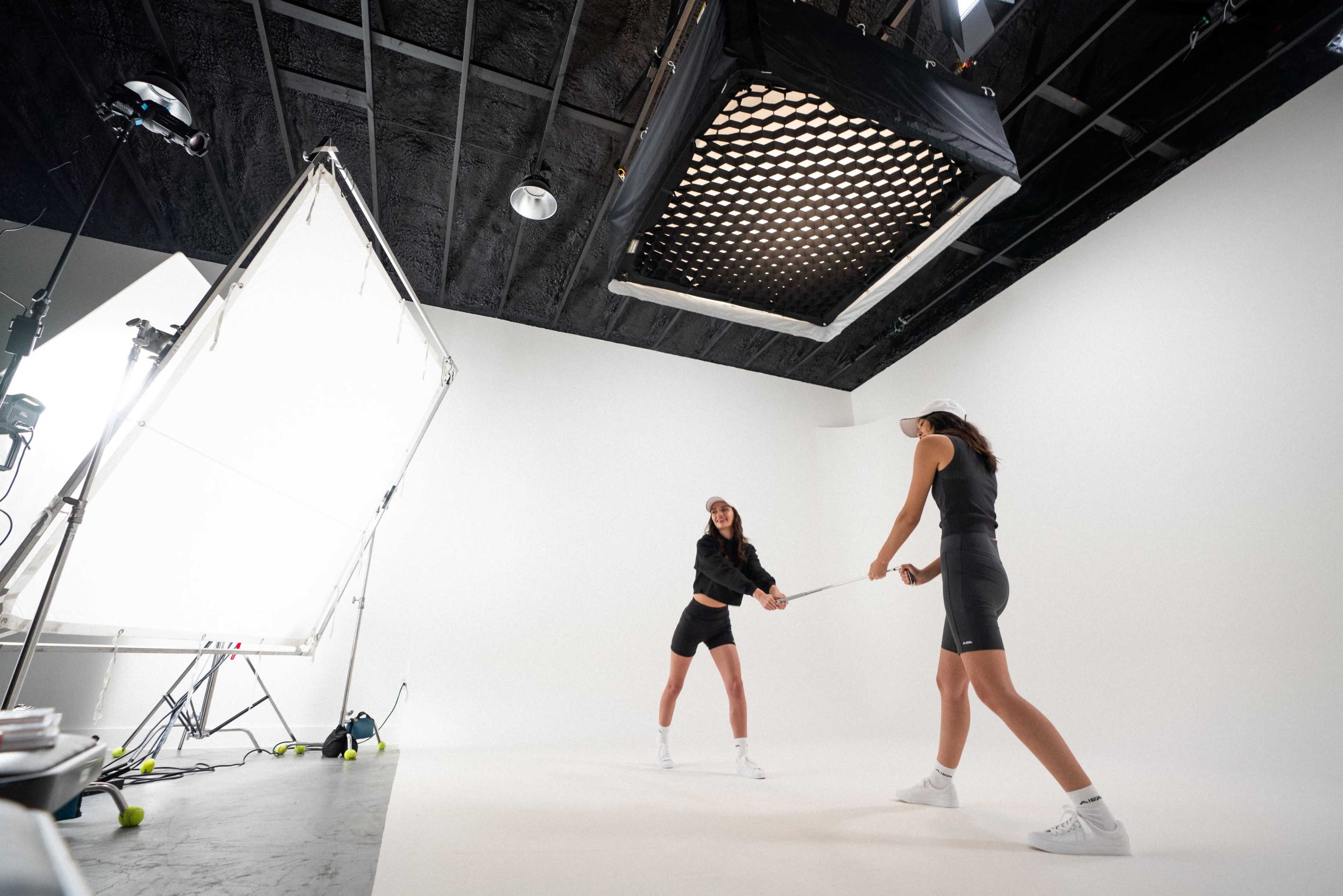 Two models are engaged in a photoshoot on a white studio backdrop, wielding rackets with bright lighting equipment above them.