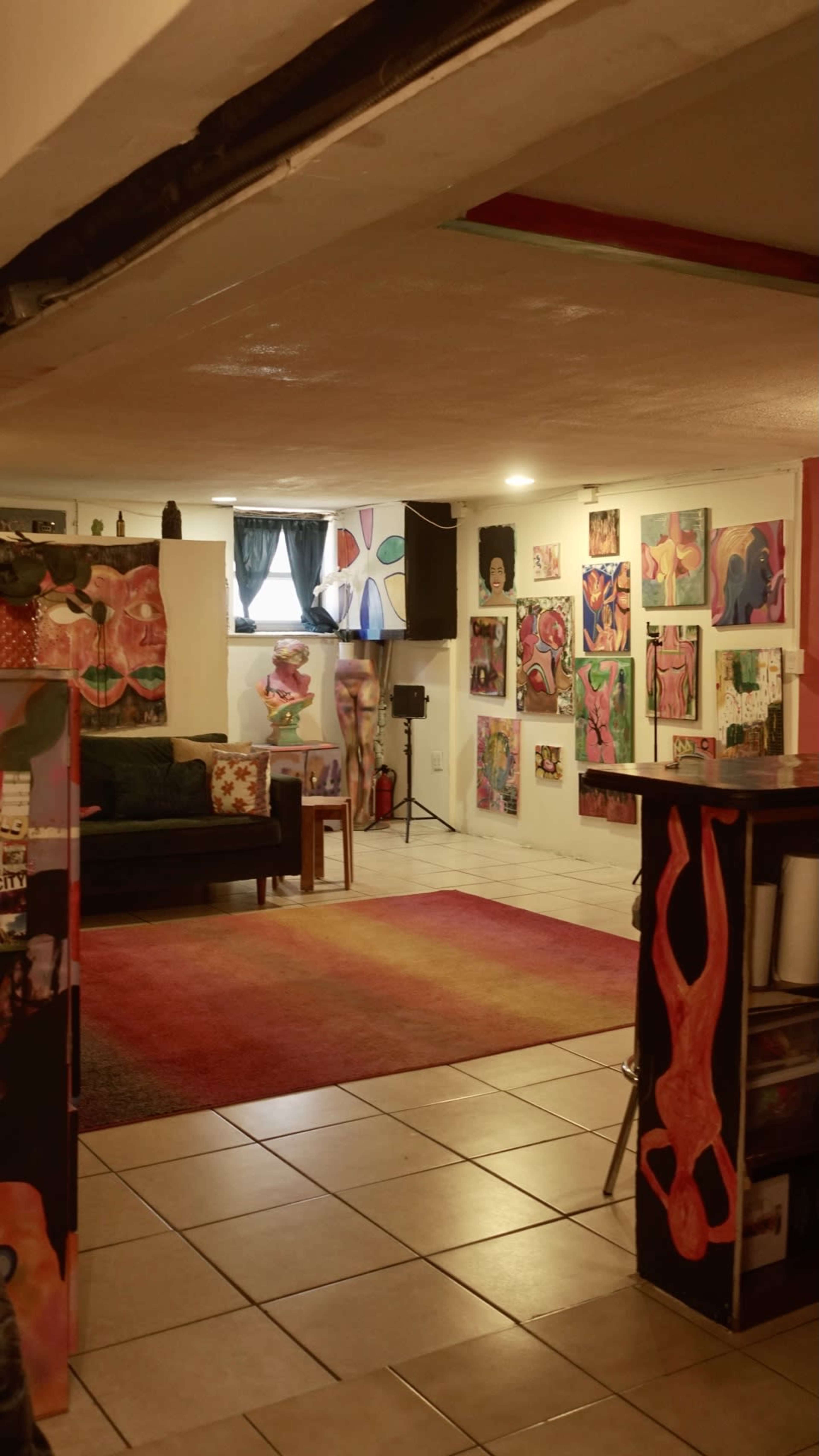 A cozy art studio or gallery featuring colorful paintings on the walls, a sofa, a rug, and a small table in a well-lit space.