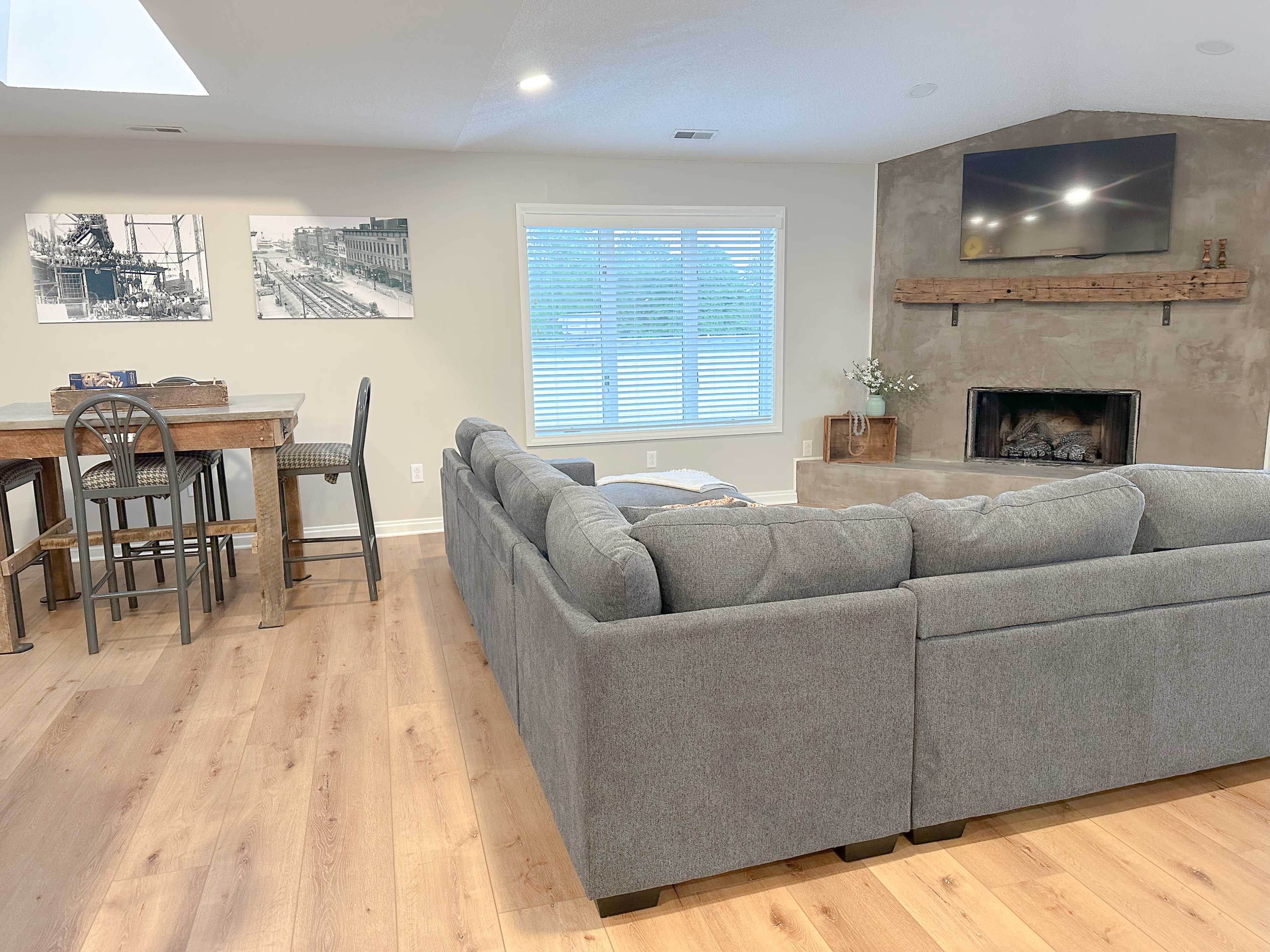 A modern living area with a gray sectional sofa, a wooden dining table and chairs, and a fireplace with a television above it.