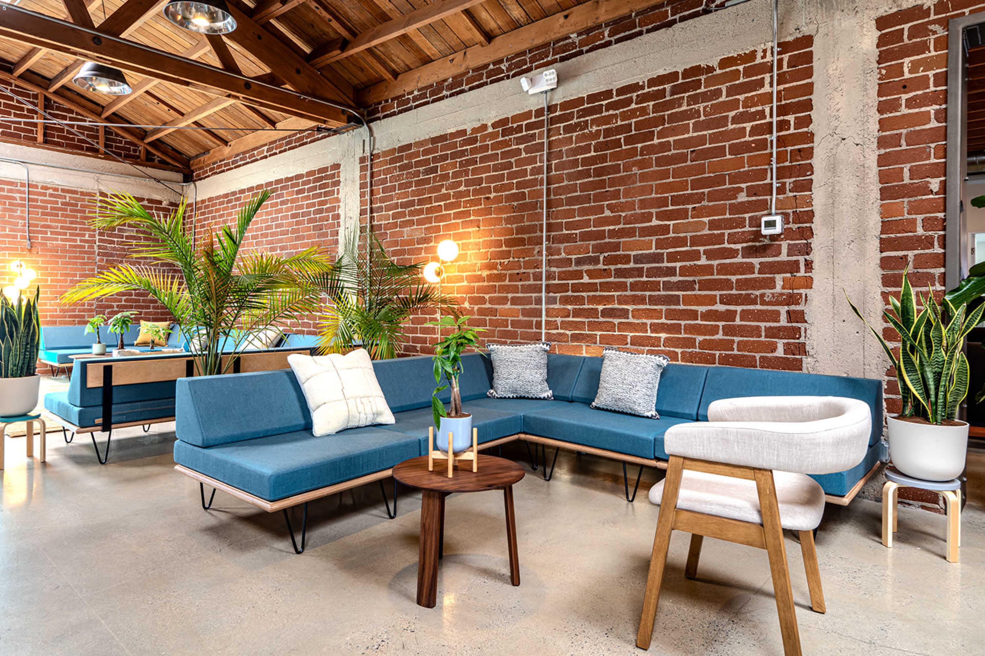 The image shows a modern lounge area featuring a blue sectional sofa, a round wooden coffee table, and potted plants against a backdrop of exposed brick walls and wooden beams.