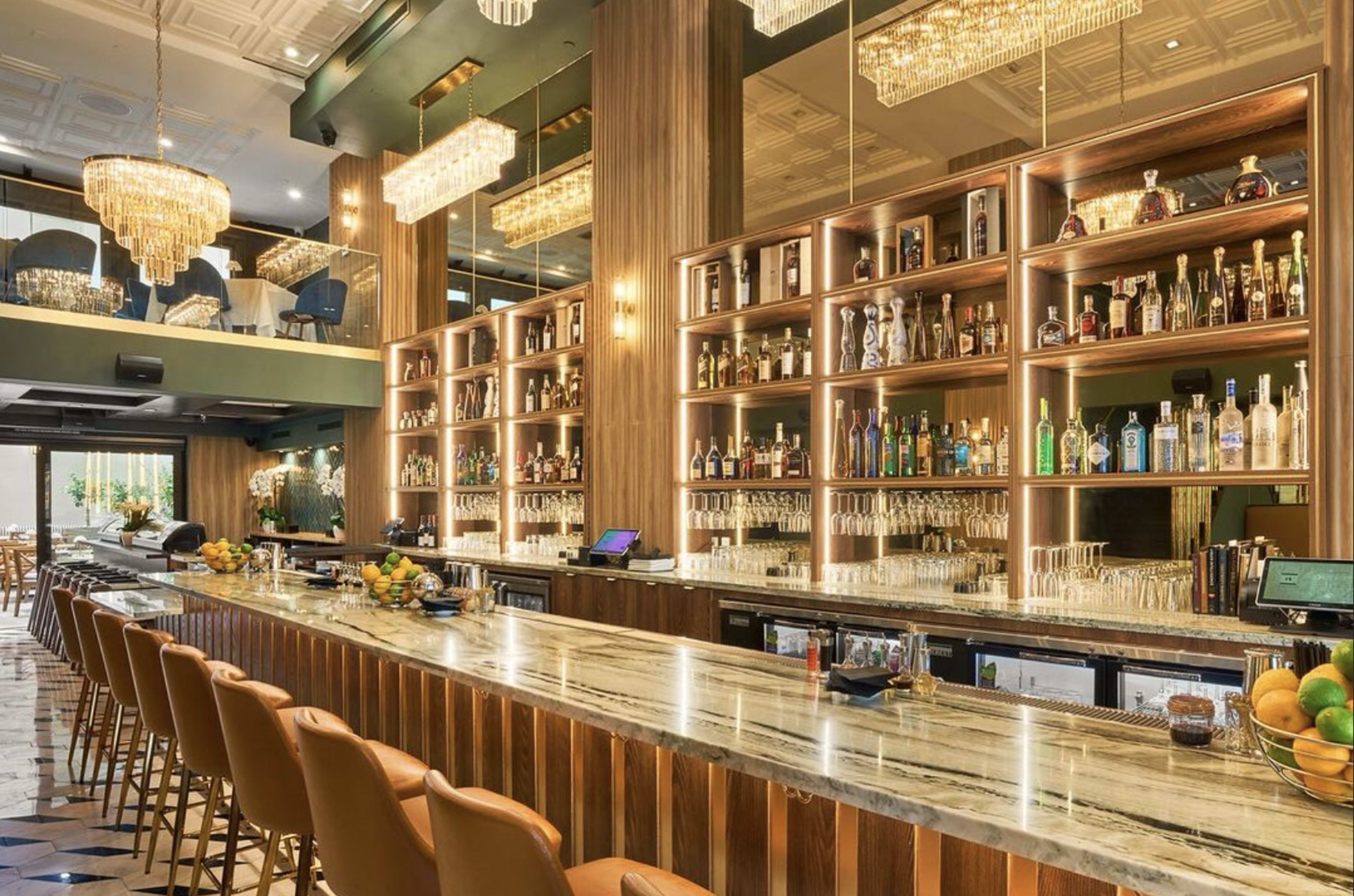 The image shows a modern bar with a marble countertop, illuminated shelves stocked with various bottles, and elegant chandeliers hanging from the ceiling.