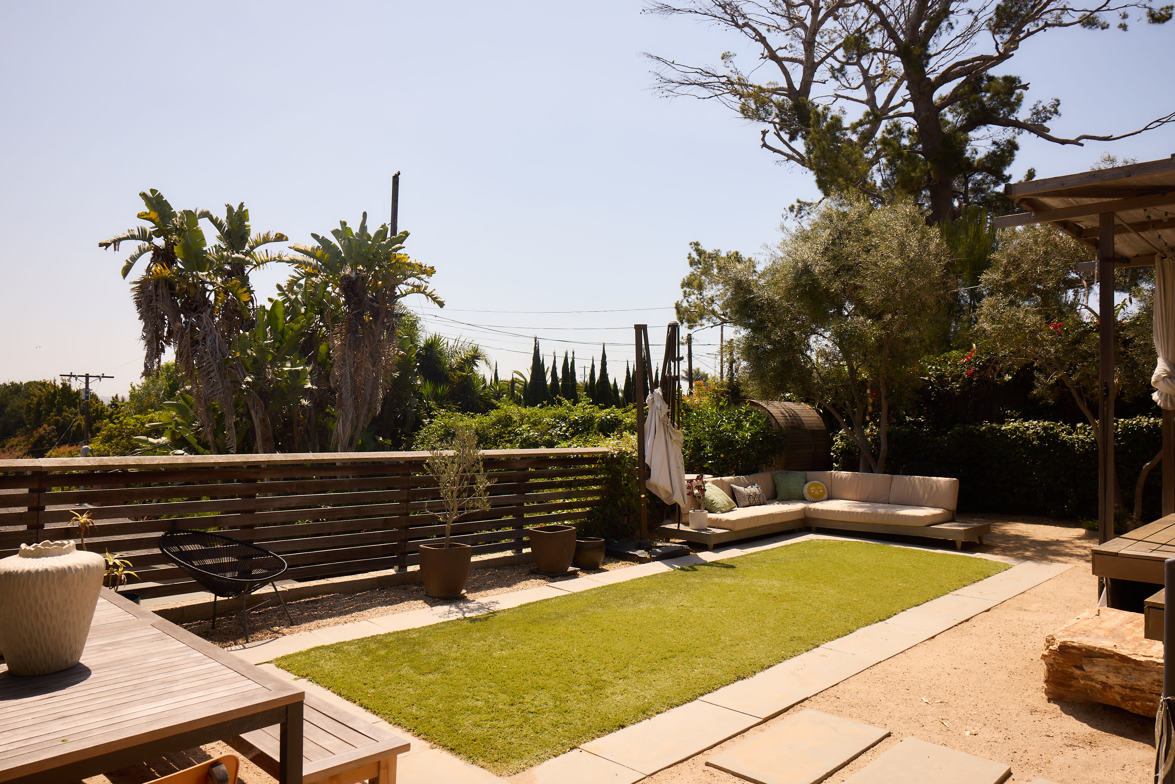 The image shows a modern outdoor patio area with a small grassy lawn, wooden deck, and potted plants surrounded by palm trees and shrubs.