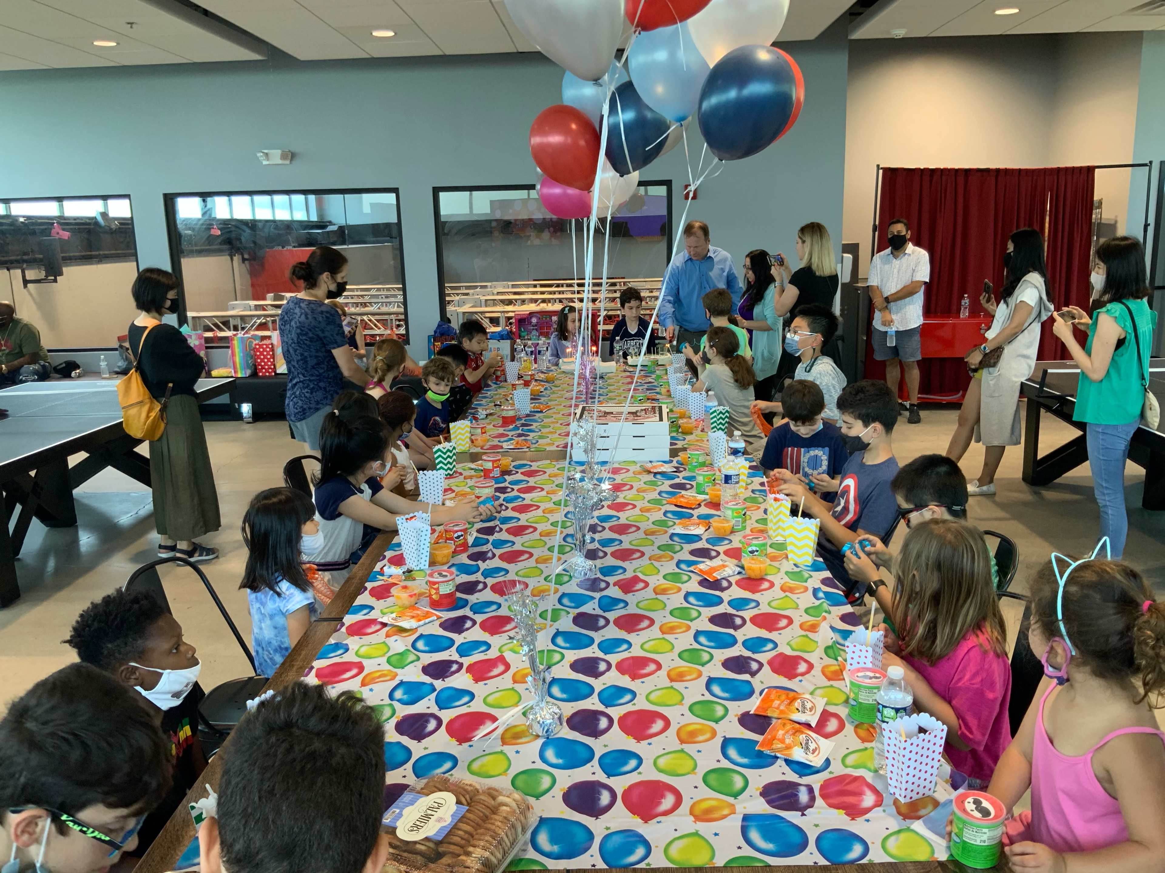 A long table covered with a colorful tablecloth is set for a gathering of children and adults, with balloons overhead and various party items arranged along the tabletop.
