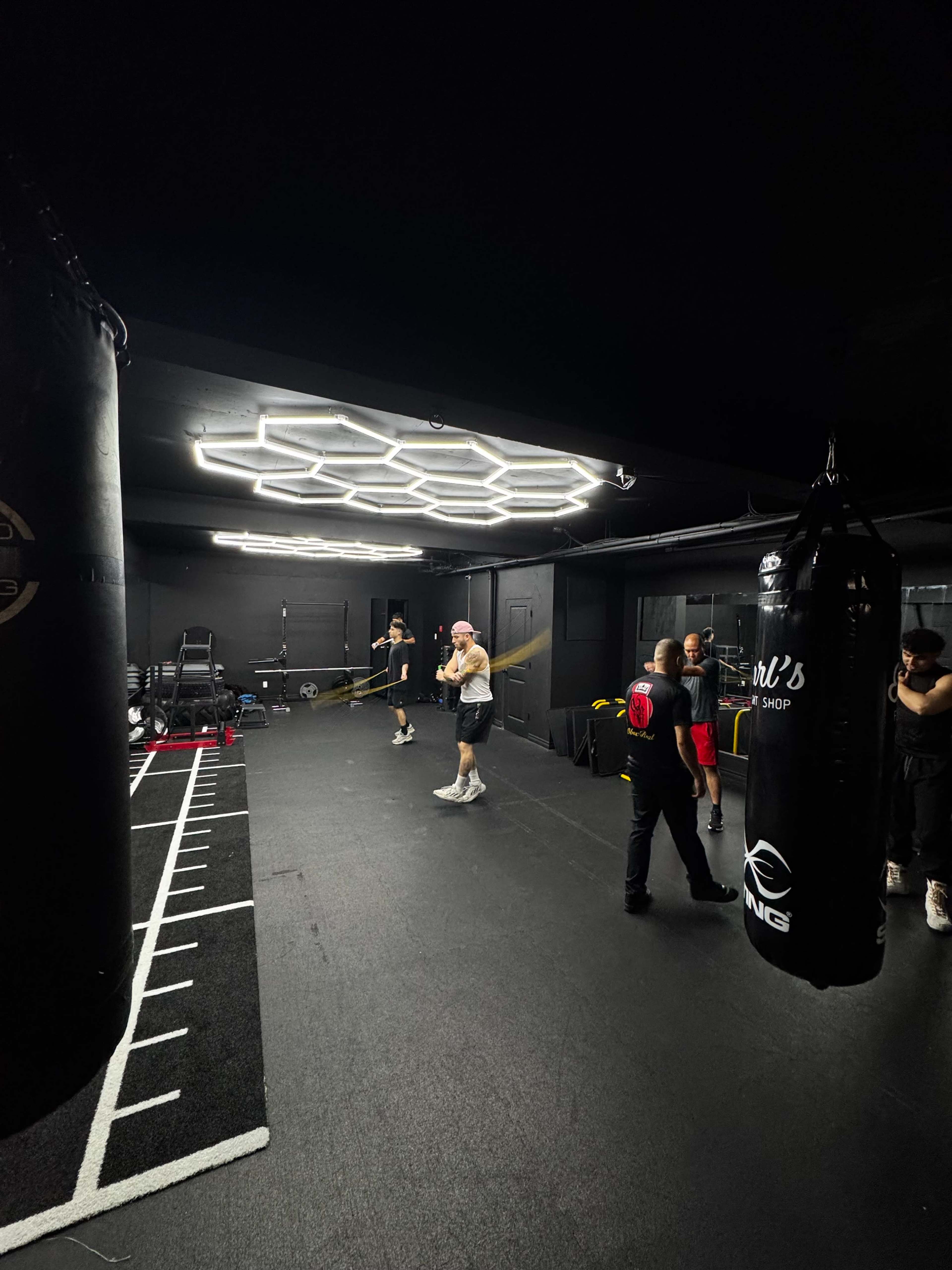 The image shows a modern boxing gym with black walls, two boxing bags, and several people engaged in various training activities.
