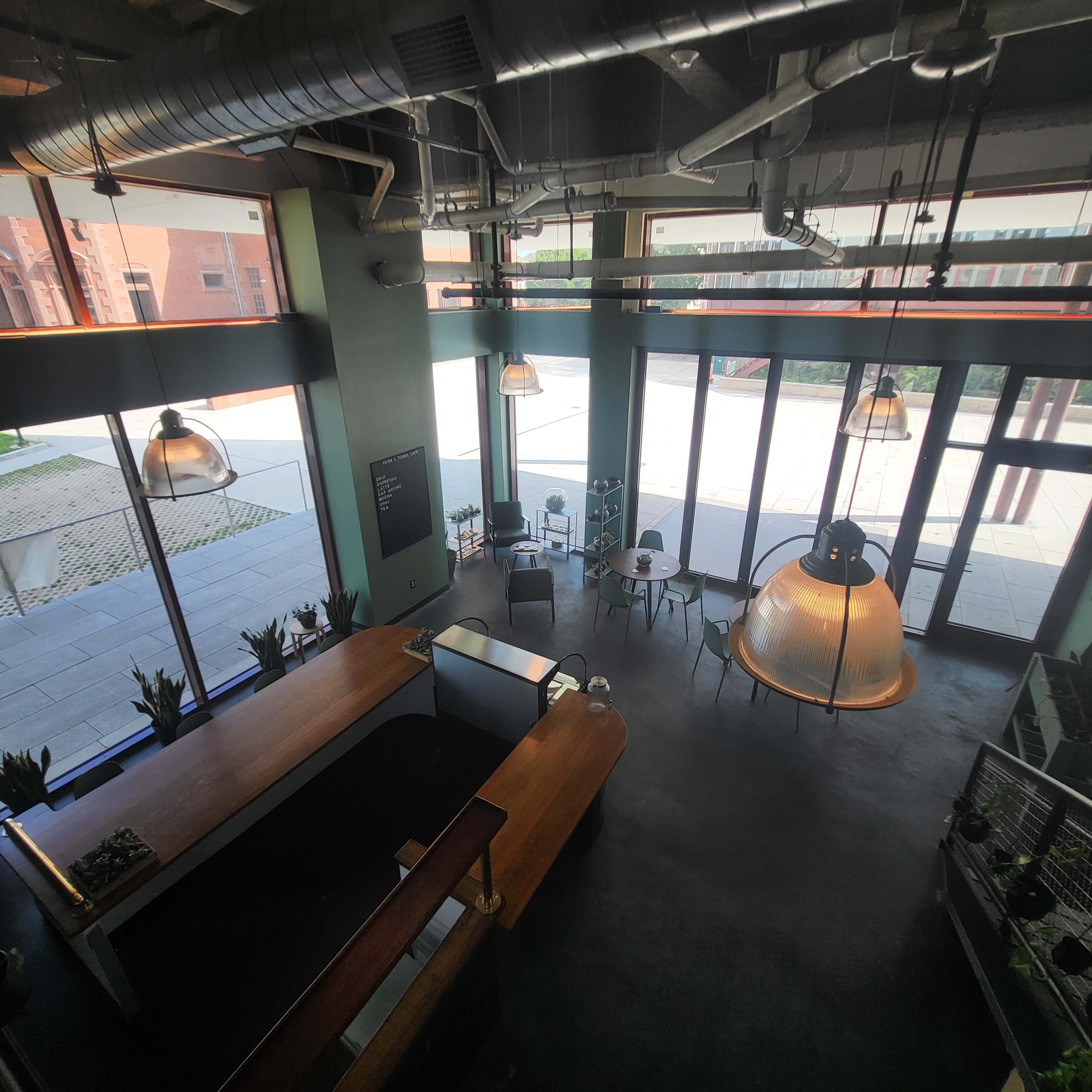 The image shows a modern café interior with large windows, hanging light fixtures, and a wooden counter, overlooking an outdoor patio area.