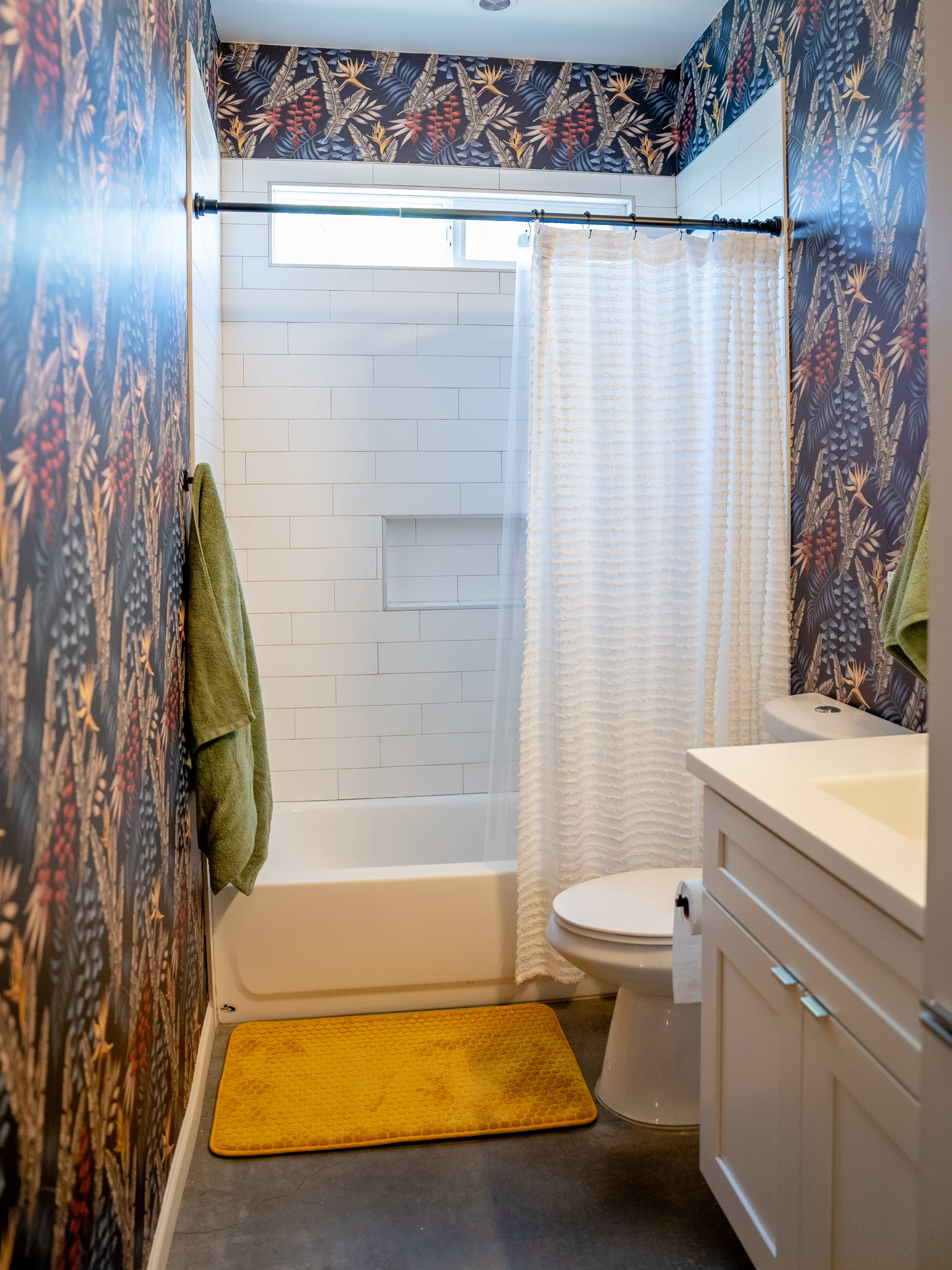 A bathroom featuring floral wallpaper, a shower curtain, and a bathtub, along with a toilet and a small yellow bath mat.
