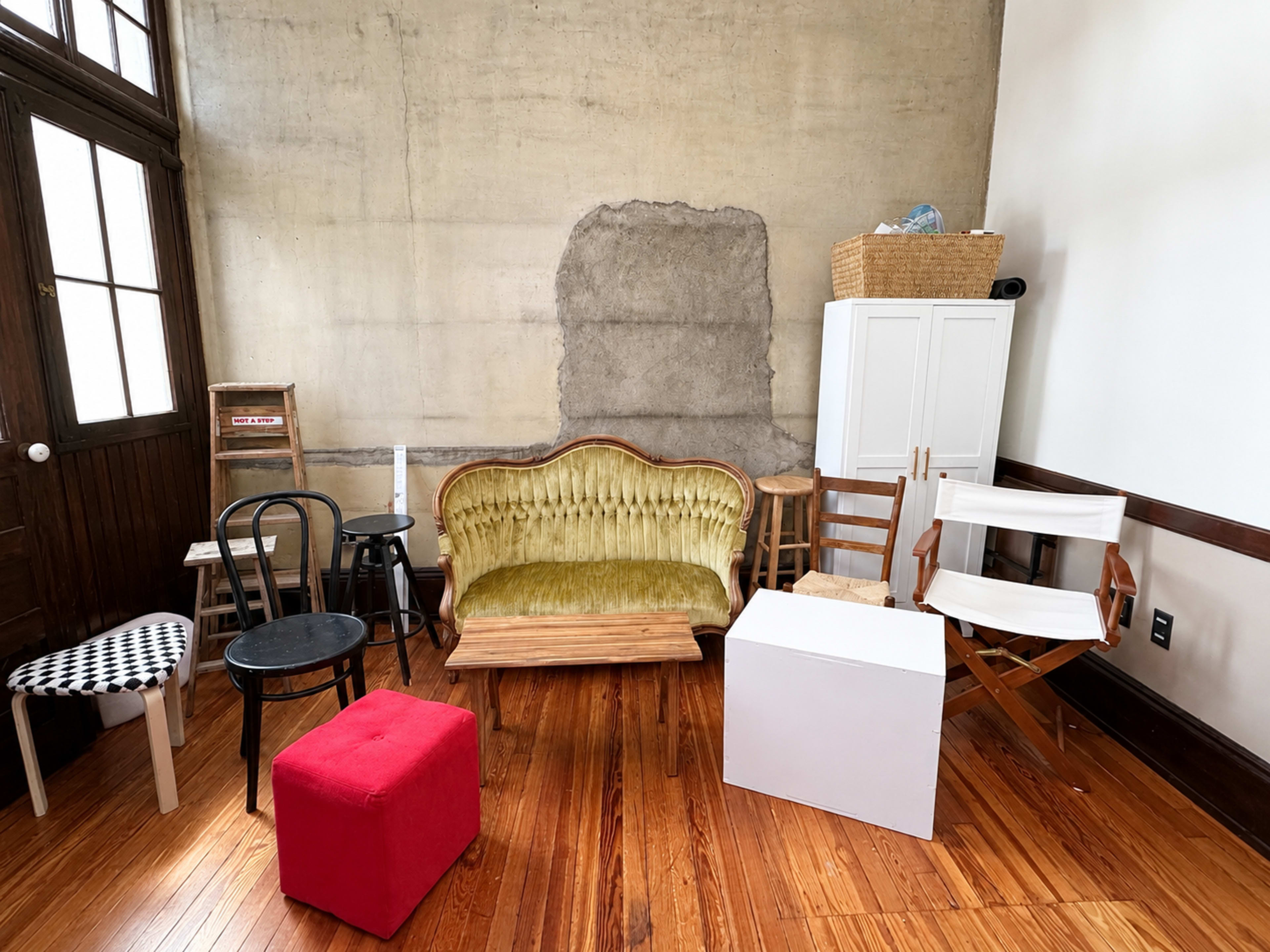 The image shows a room with a textured concrete wall, featuring various chairs, a vintage green sofa, a white cabinet, and a basket on top of it, all arranged on a wooden floor.