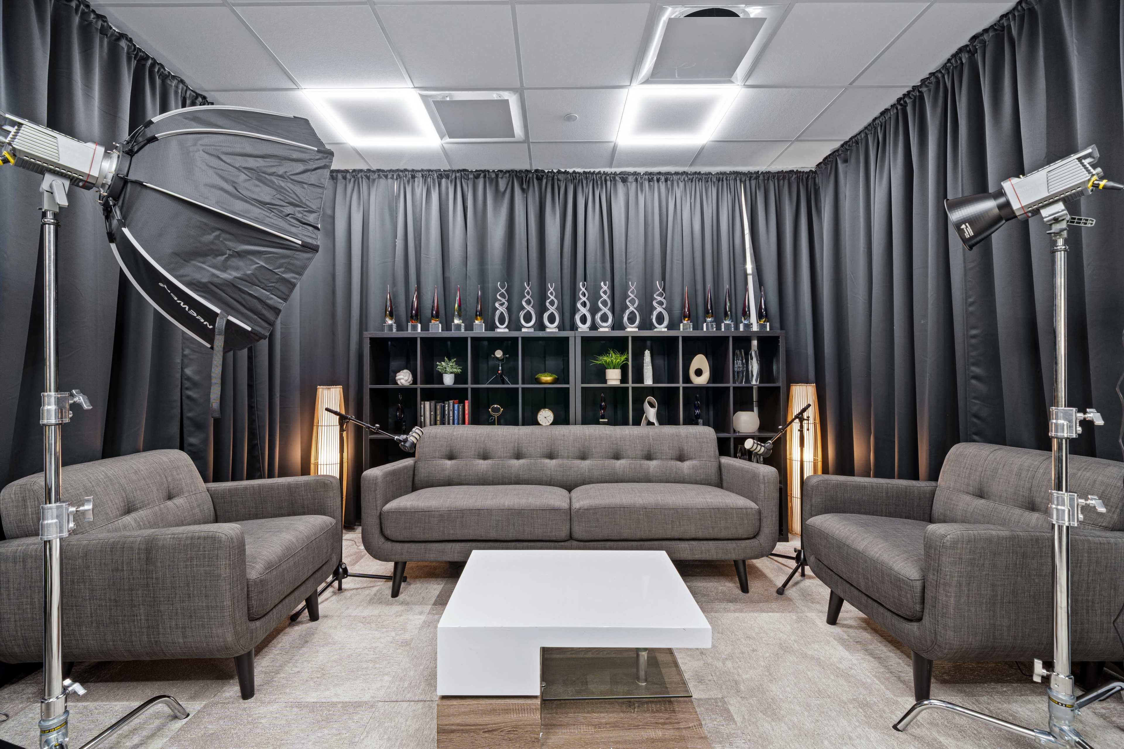 The image shows a modern studio space with gray sofas, a white coffee table, and professional lighting equipment, surrounded by dark curtains and decorative shelving.