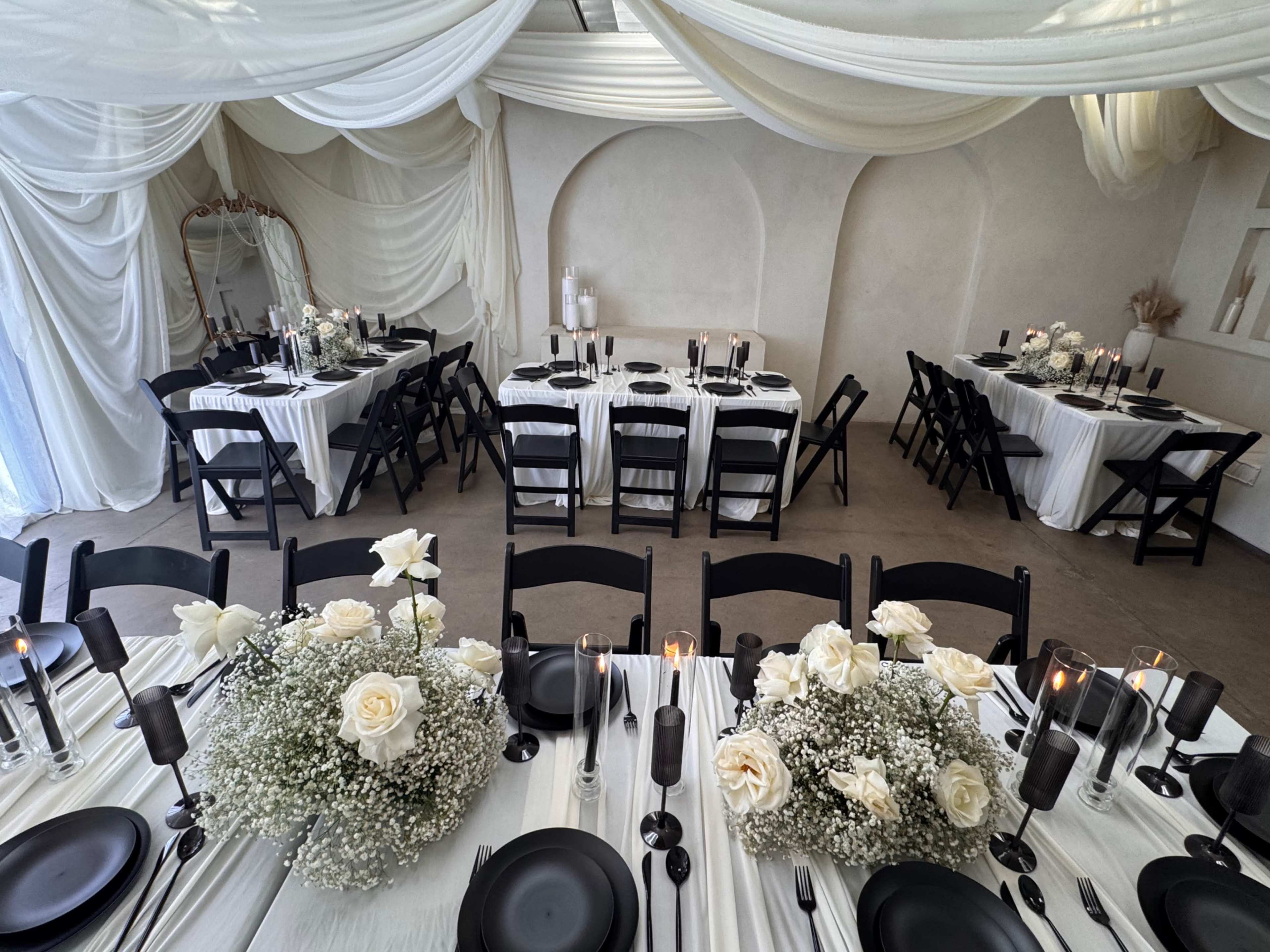 The image shows an elegantly arranged dining area with white tablecloths, black plates, and floral centerpieces featuring white roses and baby's breath under draped fabric decor.