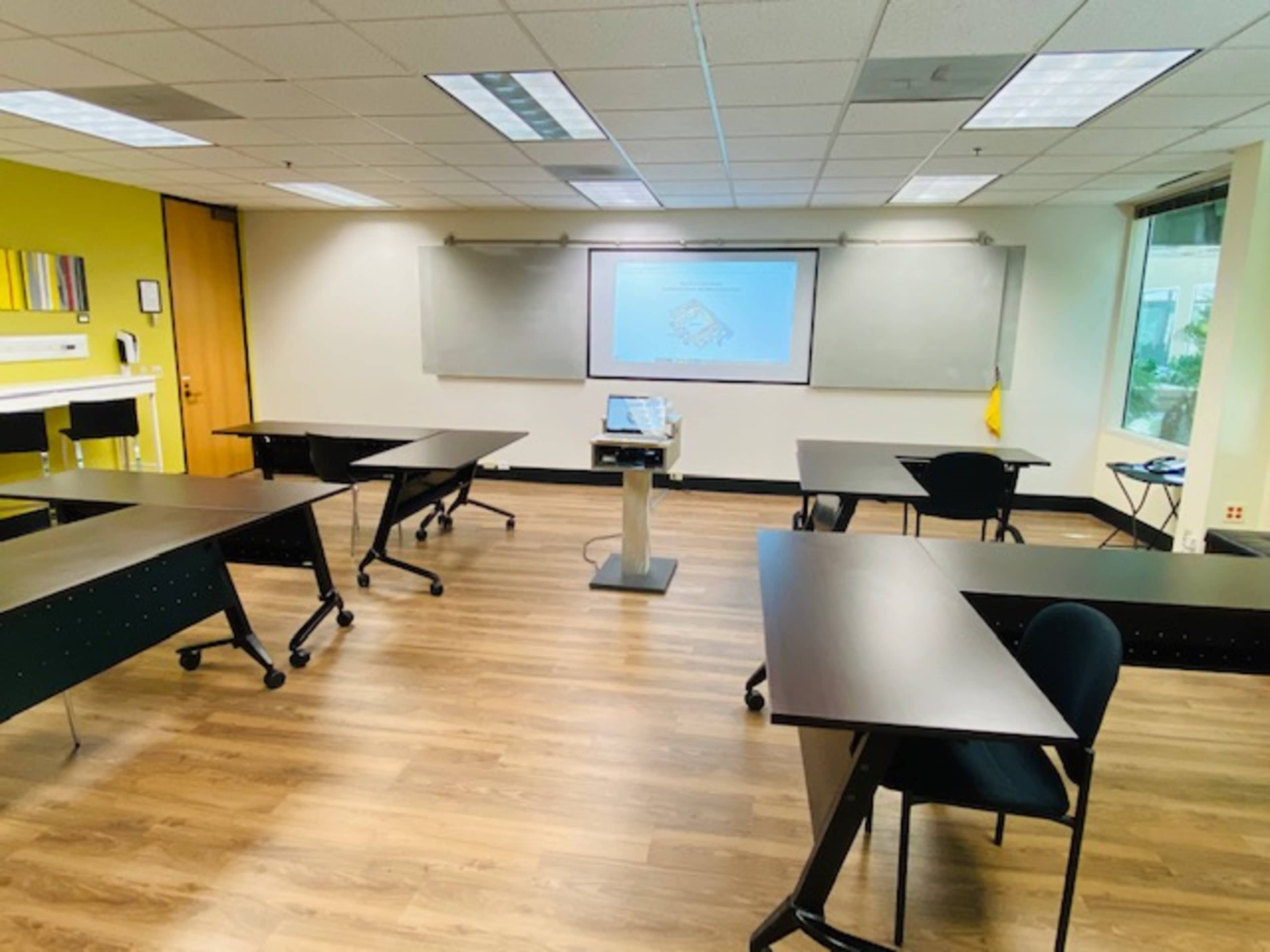 A classroom set up with multiple tables and chairs arranged around a projector screen.