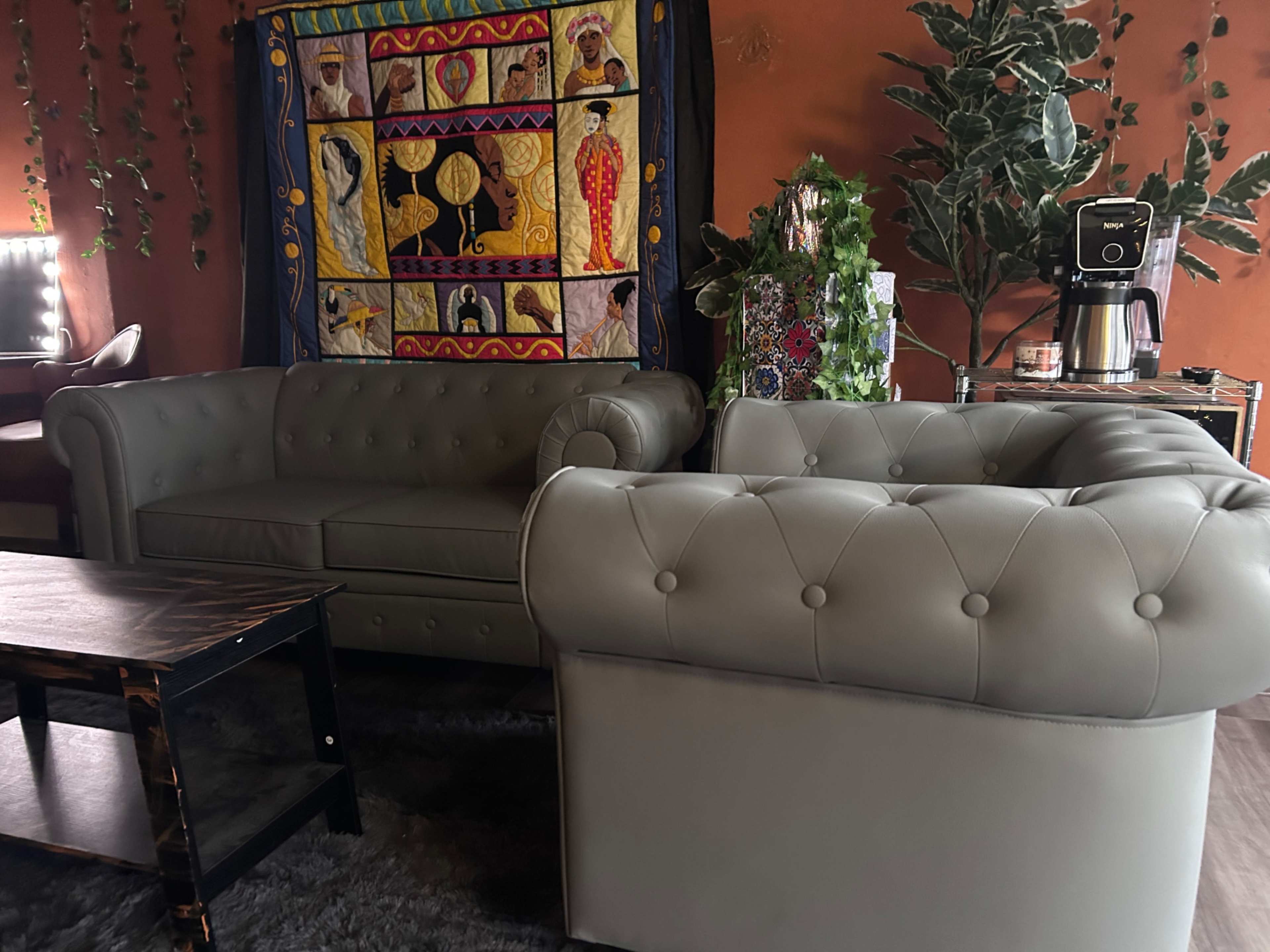The image shows two gray tufted sofas arranged near a wooden coffee table, with a colorful wall hanging and a coffee machine in the background.