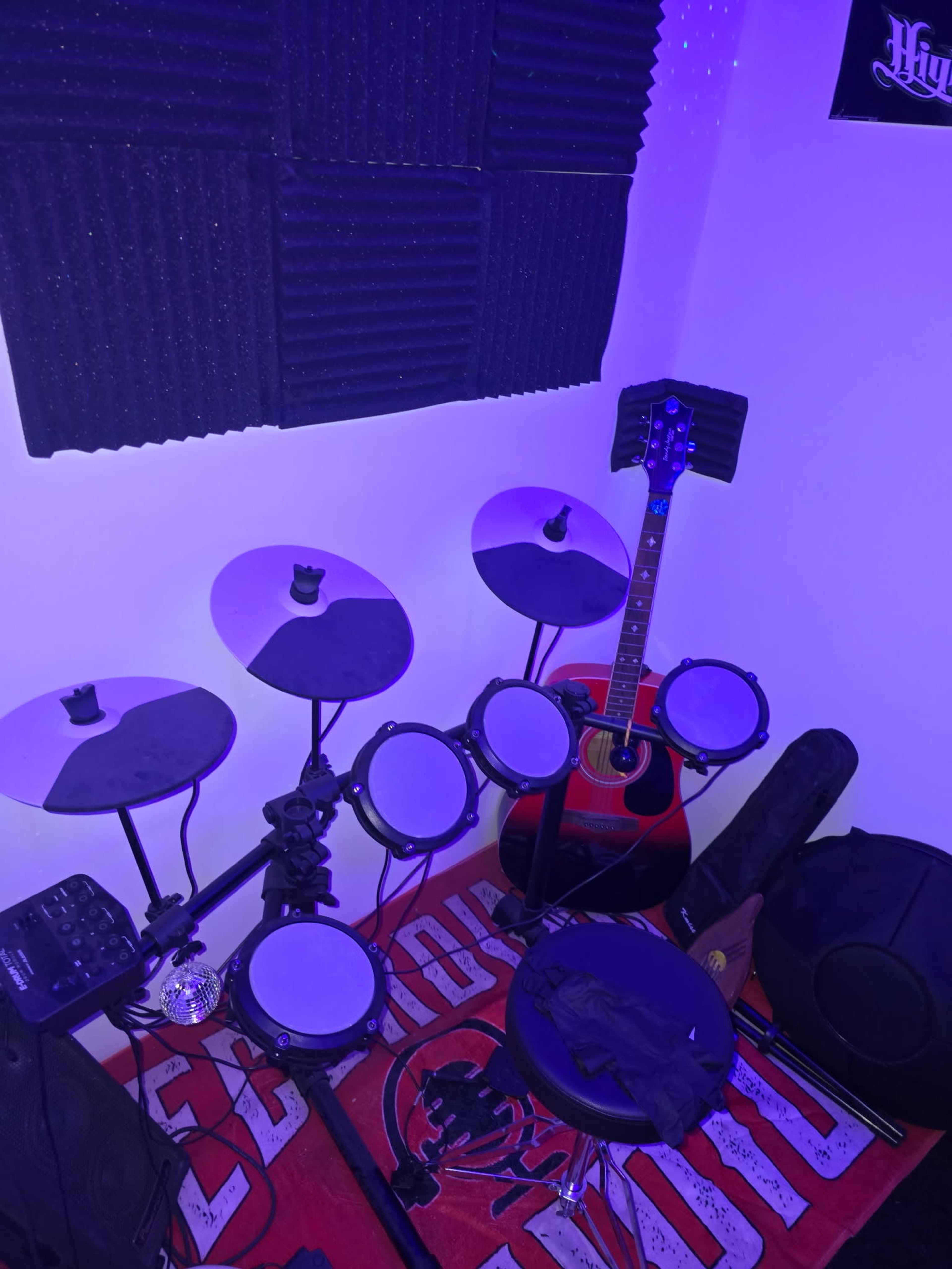 The image shows an electronic drum set next to an acoustic guitar in a small, dimly lit music practice space.
