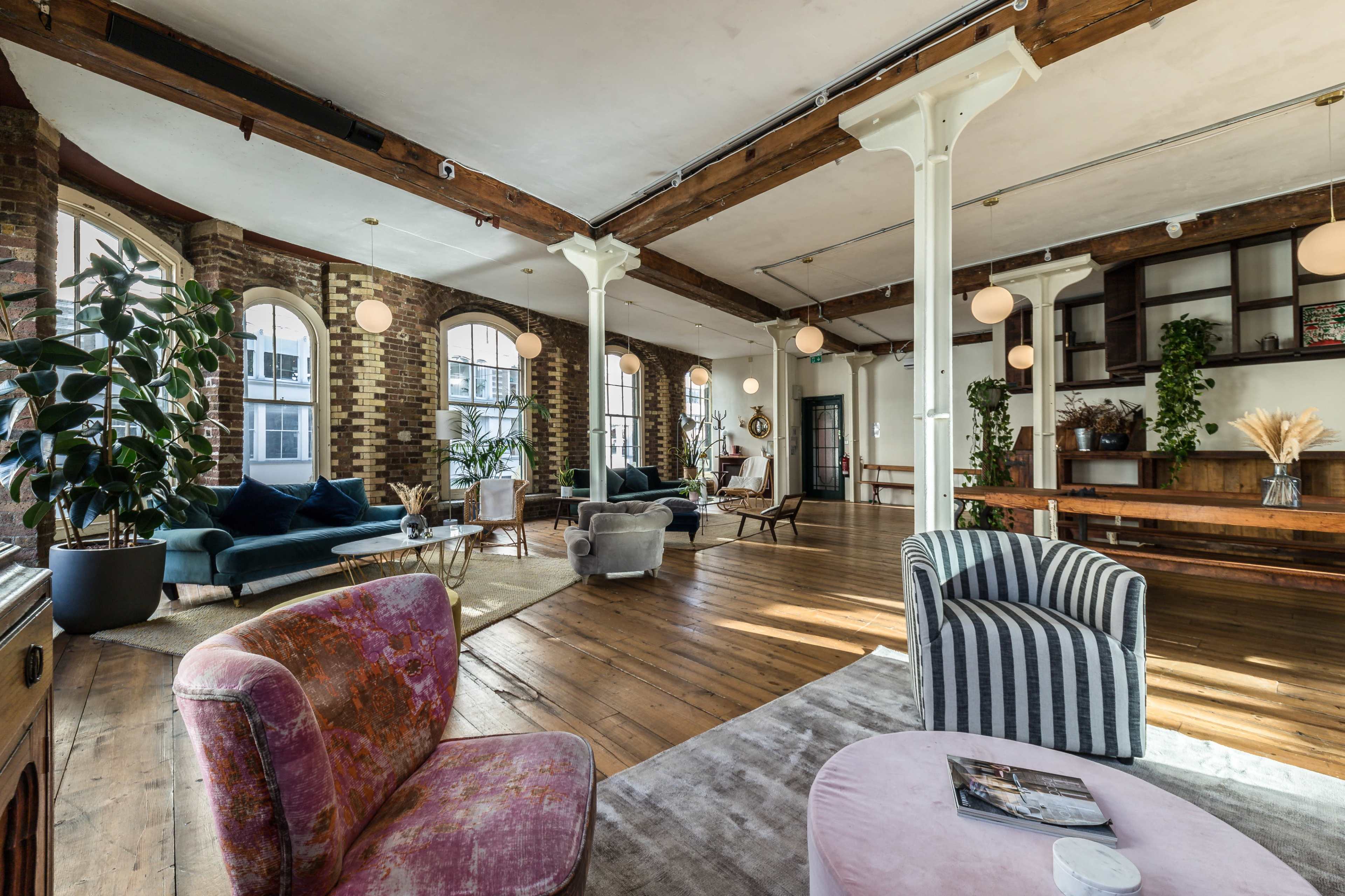 A spacious, well-lit interior with exposed brick walls, wooden beams, and various seating areas arranged around a central wooden floor.