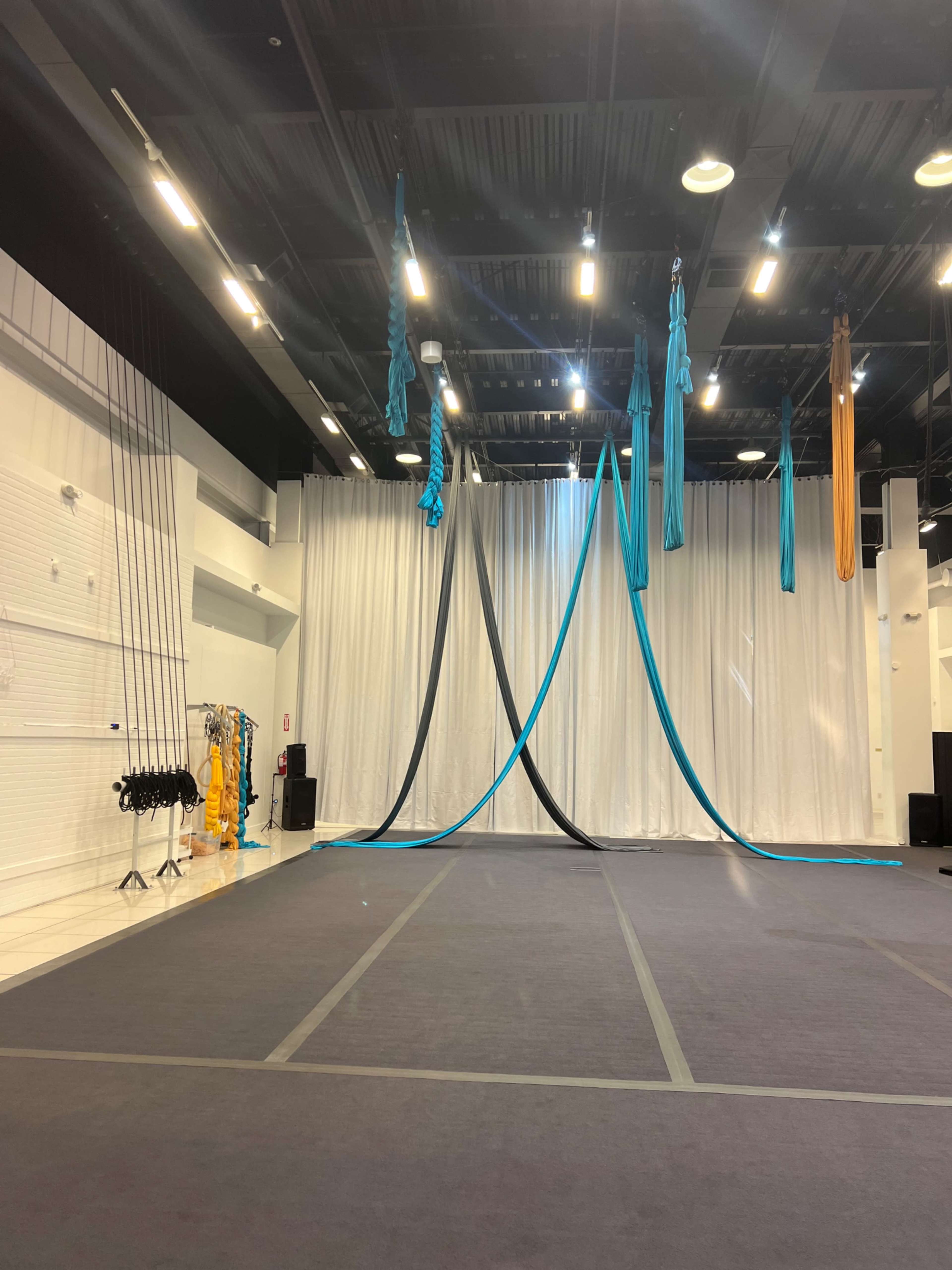 The image shows a spacious studio with dark flooring, a white backdrop, and aerial silk fabric hanging from the ceiling in various colors.