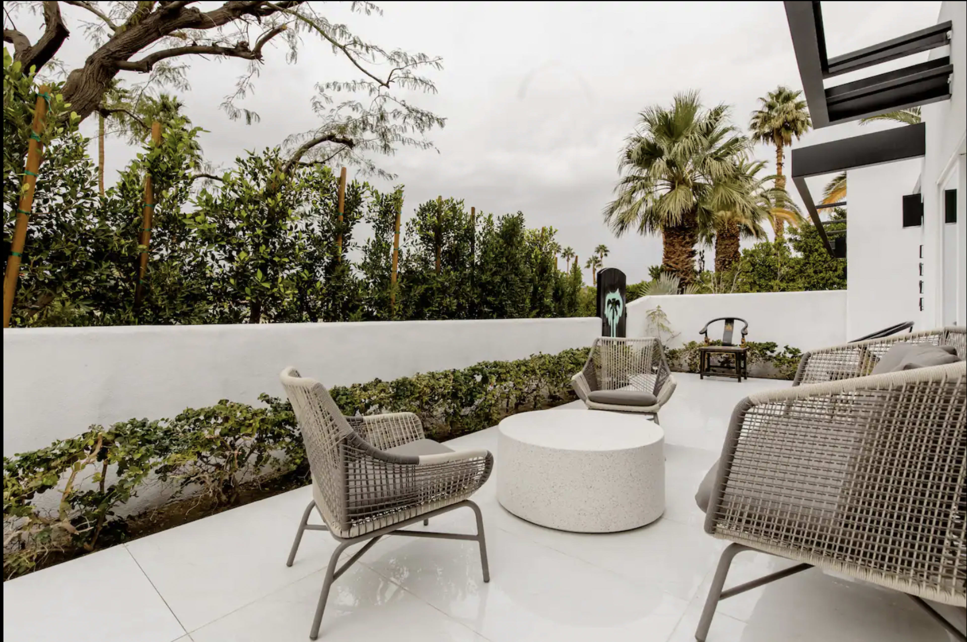 A patio features several modern chairs arranged around a round table, with lush greenery and palm trees in the background.