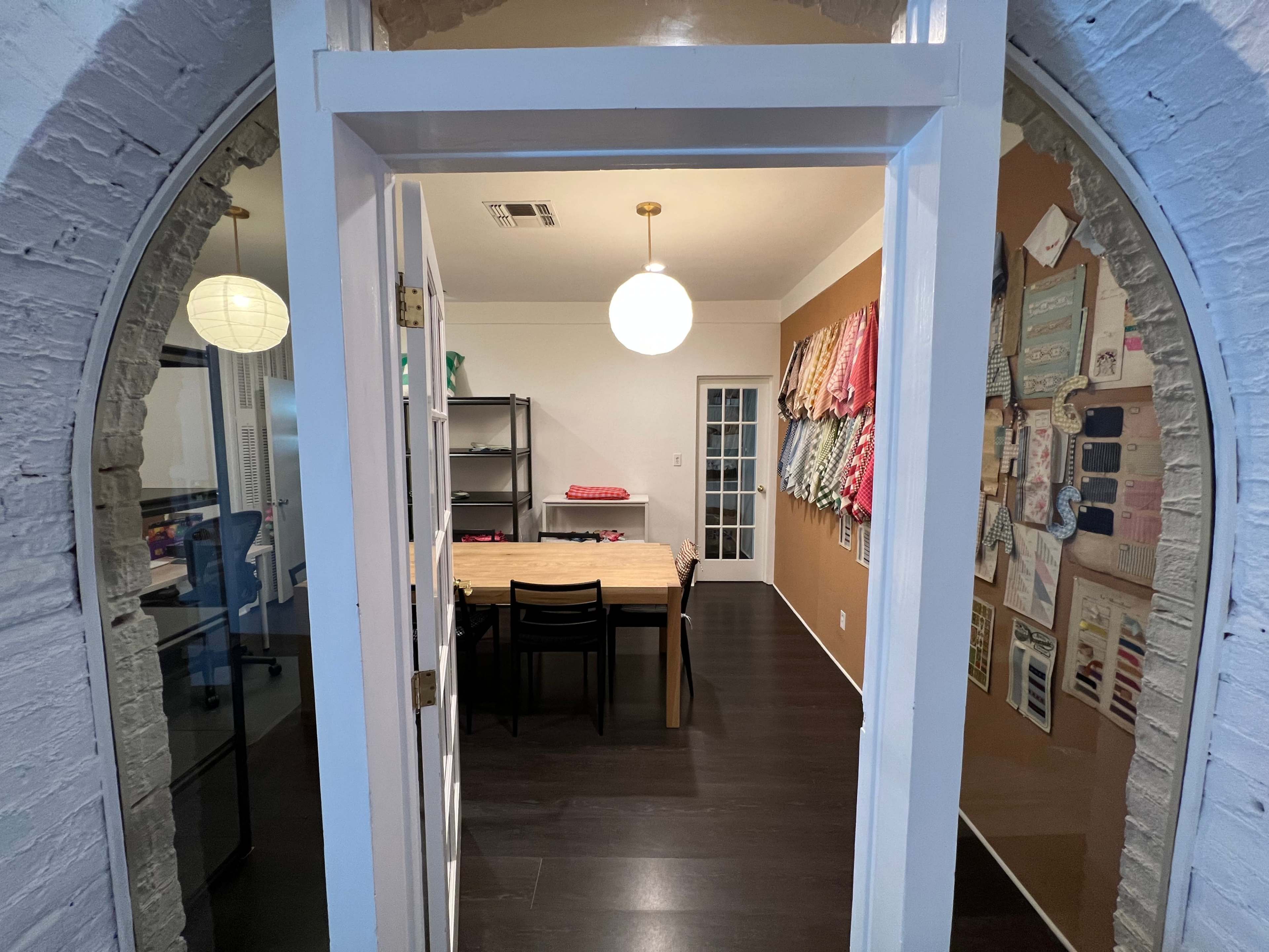 The image shows a well-lit room with a central wooden table, black chairs, and fabric samples displayed on the wall.