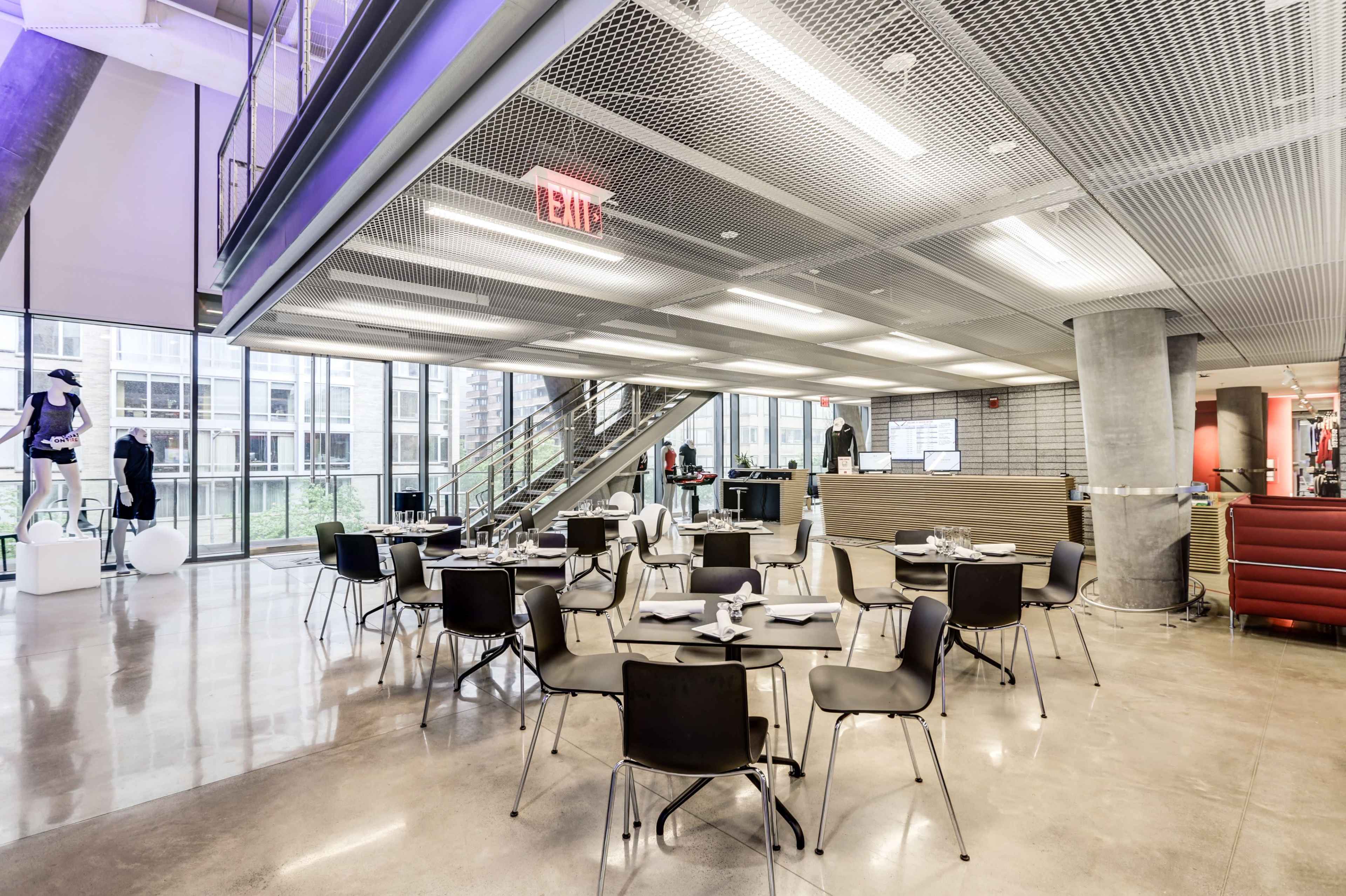 The image shows a modern dining area with several round tables and black chairs, featuring large windows that allow natural light to enter the space.