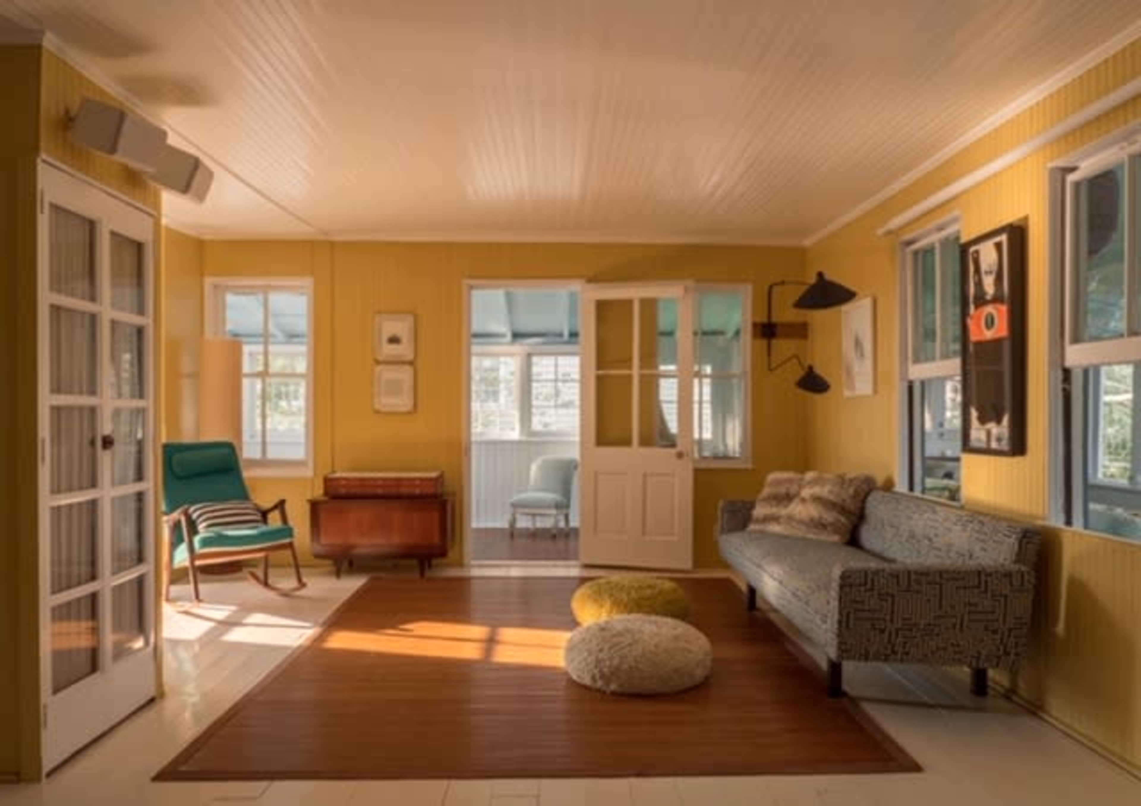 The image shows a bright, yellow-walled room featuring a sofa, armchair, a wooden side table, and large windows letting in natural light.