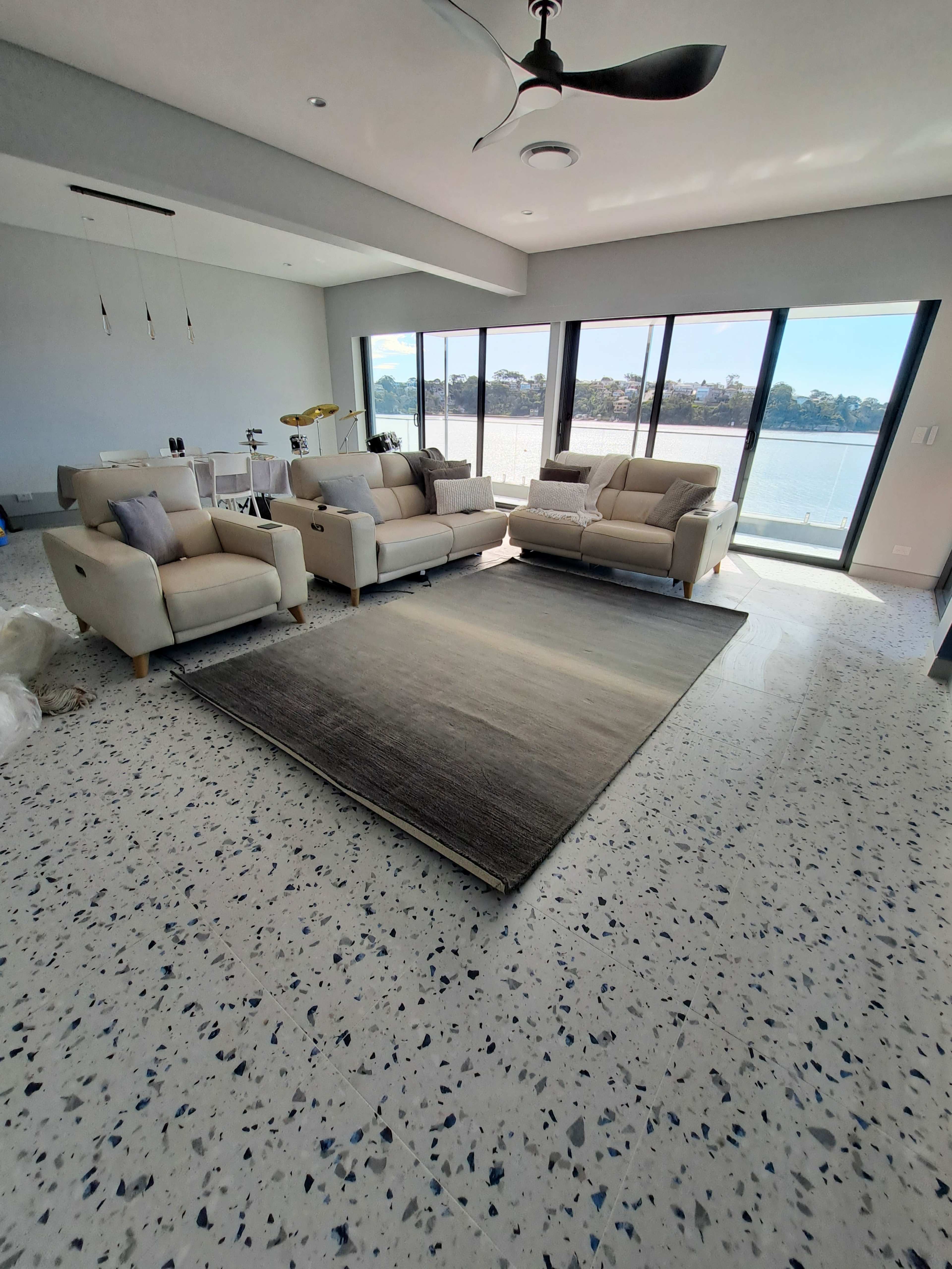 The image shows a spacious living room with two light-colored sofas, a large area rug, and floor-to-ceiling windows overlooking a waterfront view.