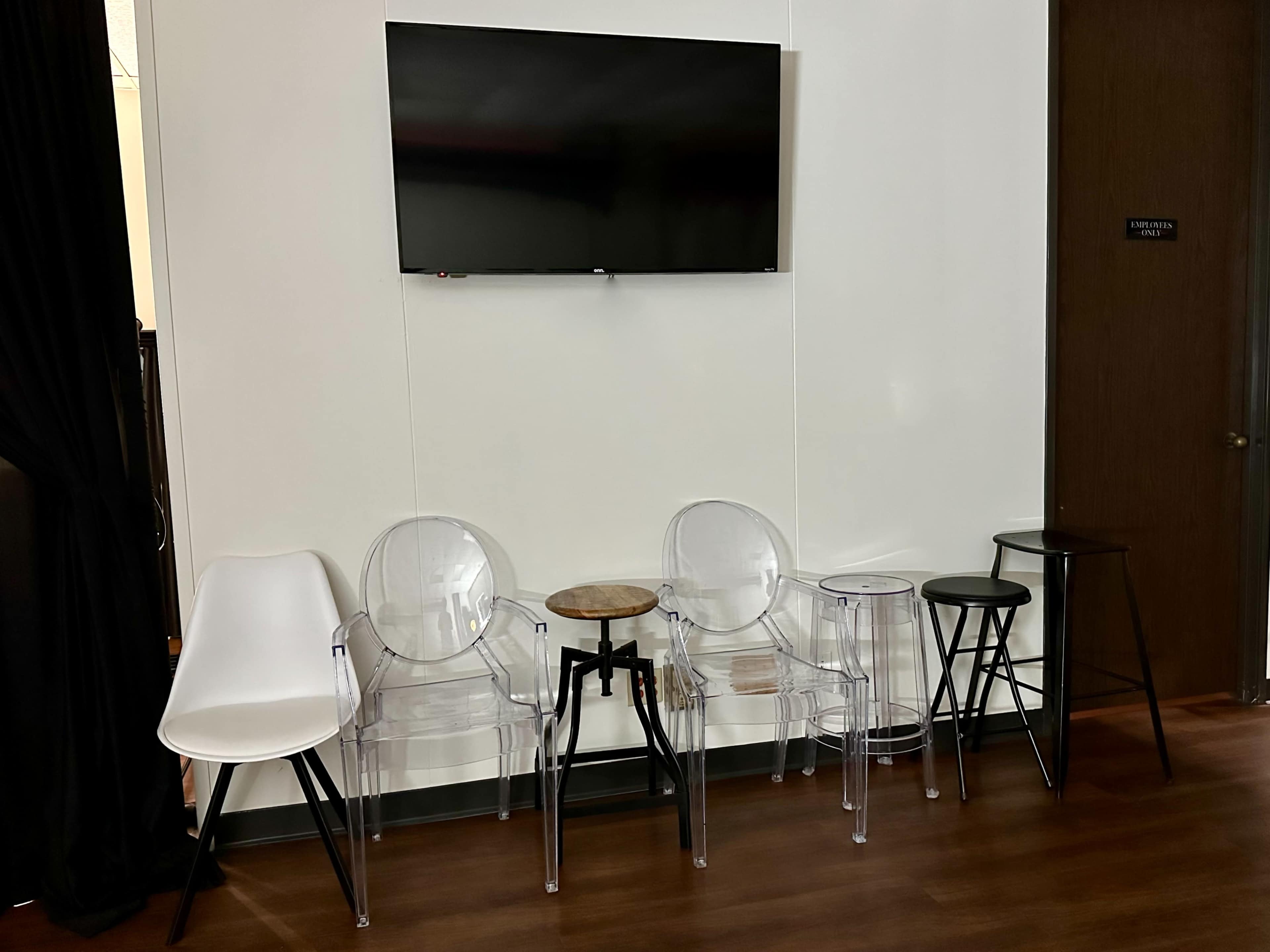 A small seating area with several transparent and solid chairs arranged next to a wooden side table and a television mounted on the wall.
