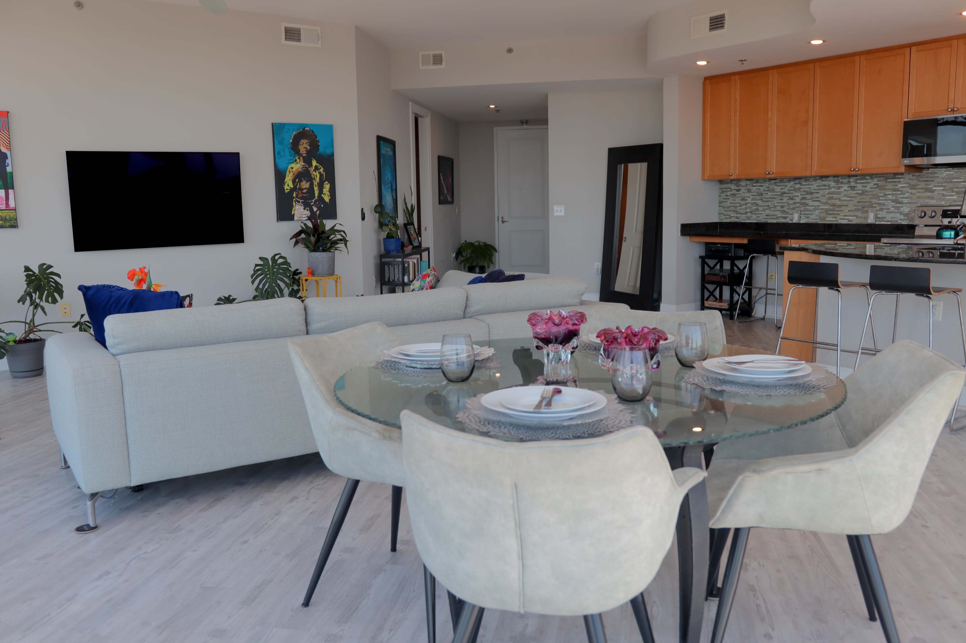 A modern living space features a dining area with a circular glass table set for a meal, surrounded by light-colored chairs, and an open lounge with a sectional sofa and wall-mounted television.