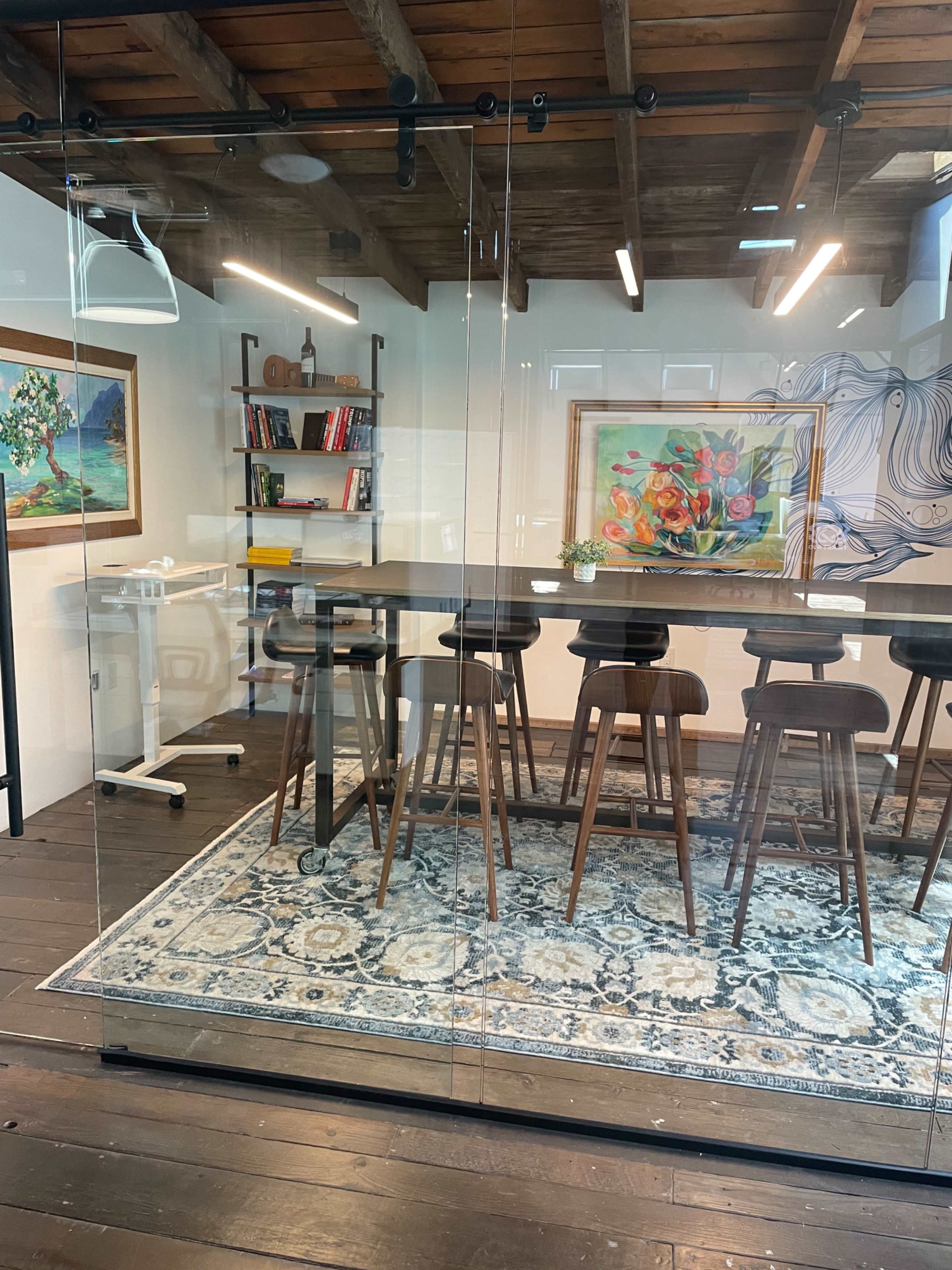 The image shows a modern office meeting room with a long table, high-backed stools, and decorative artwork on the walls, all enclosed by glass walls.