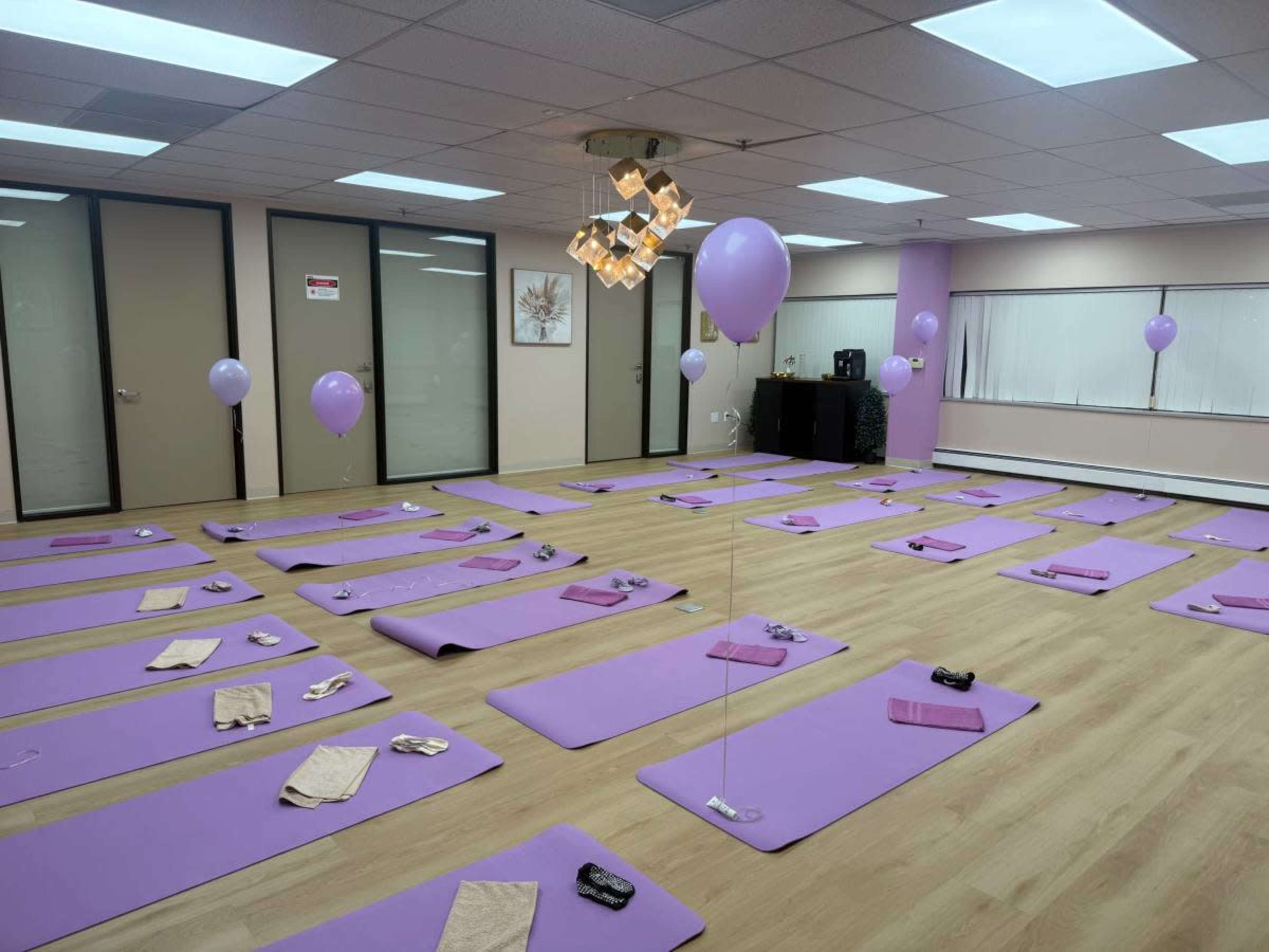 A spacious yoga studio is set up with purple mats laid out in rows, adorned with balloons and small items on each mat.
