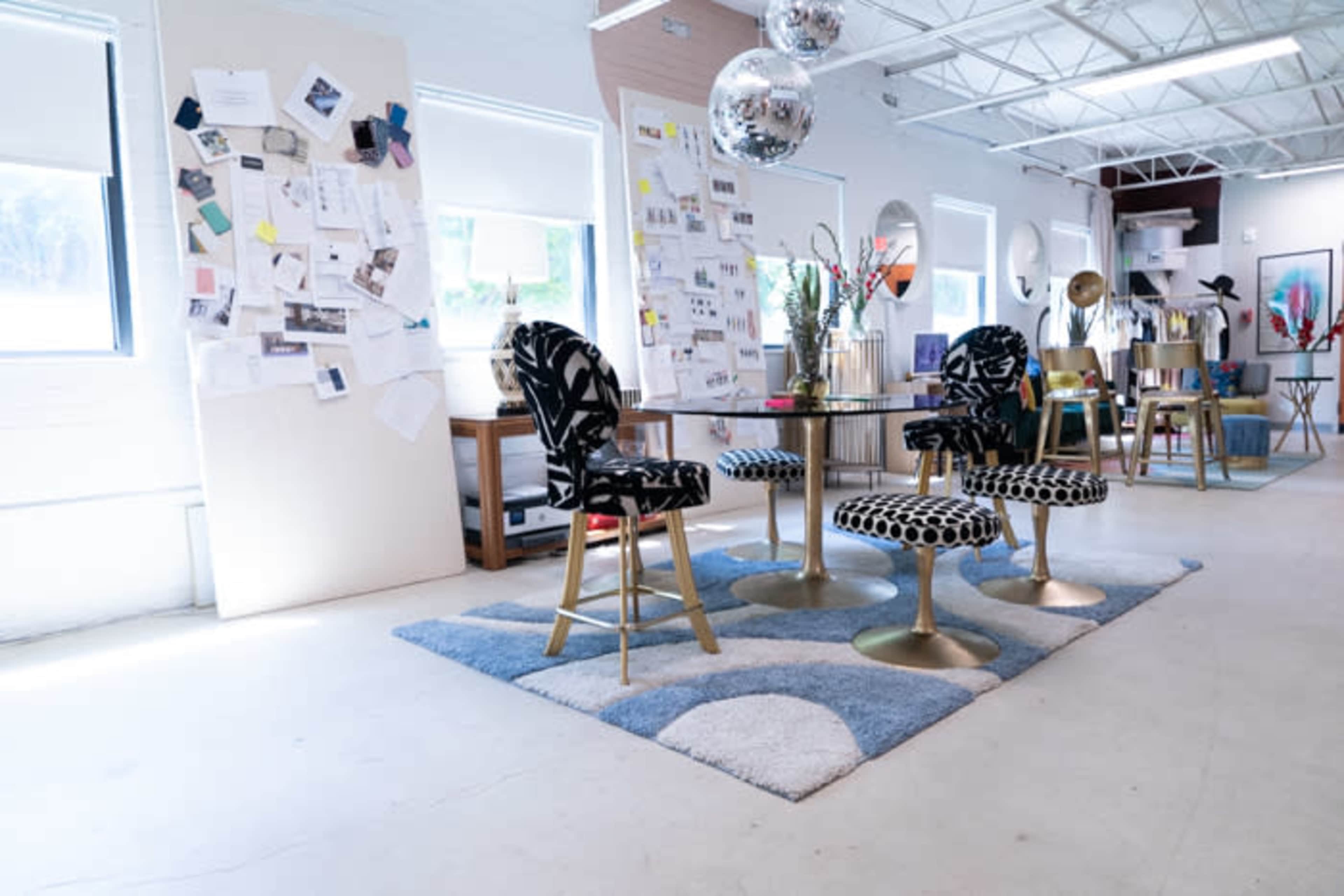 A bright interior space featuring a circular table with four upholstered chairs, a blue patterned rug, and various design materials pinned to the wall.