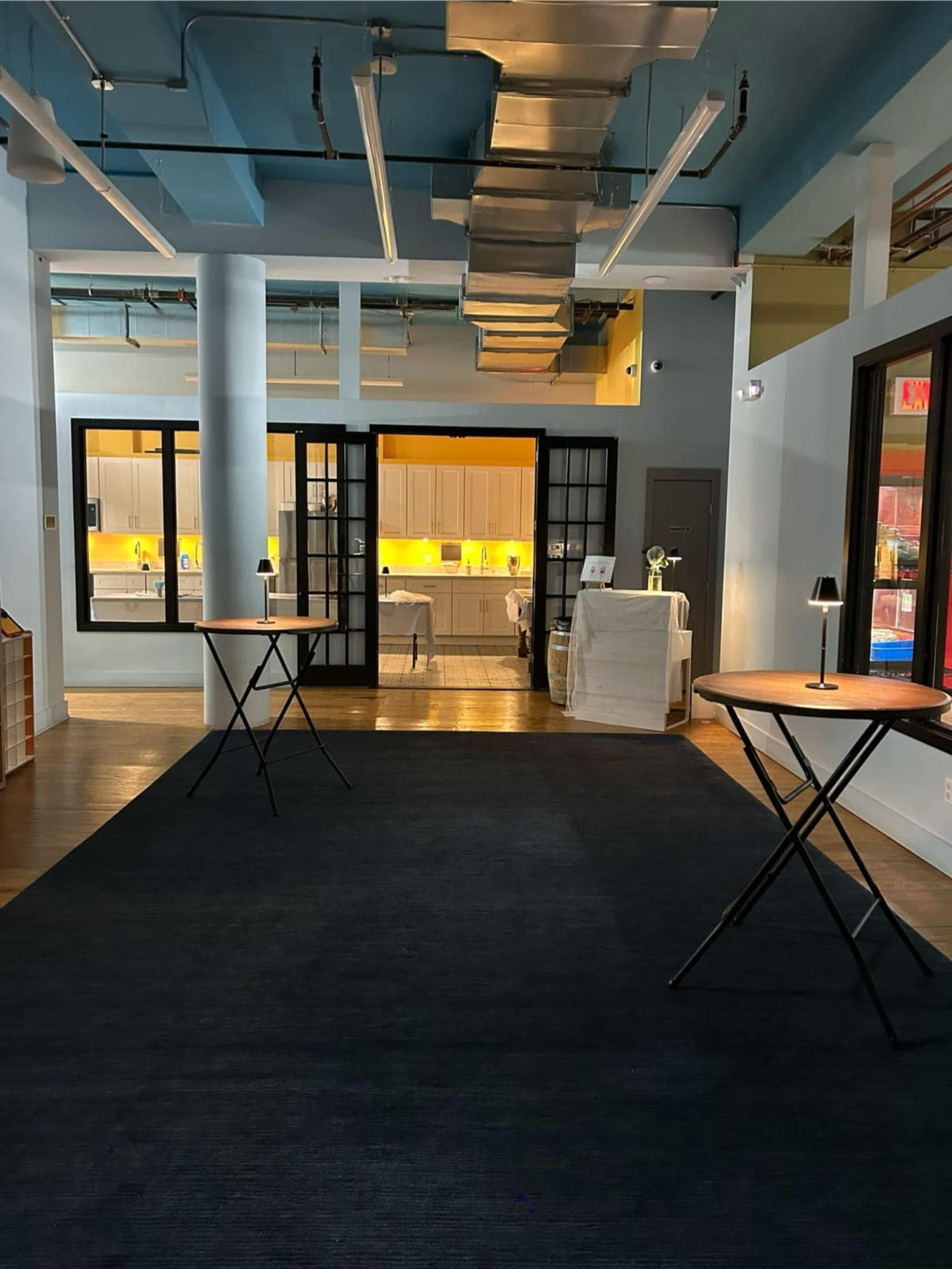 The image shows a spacious indoor area with two round tables in the foreground and a well-lit kitchen visible through double doors in the background.