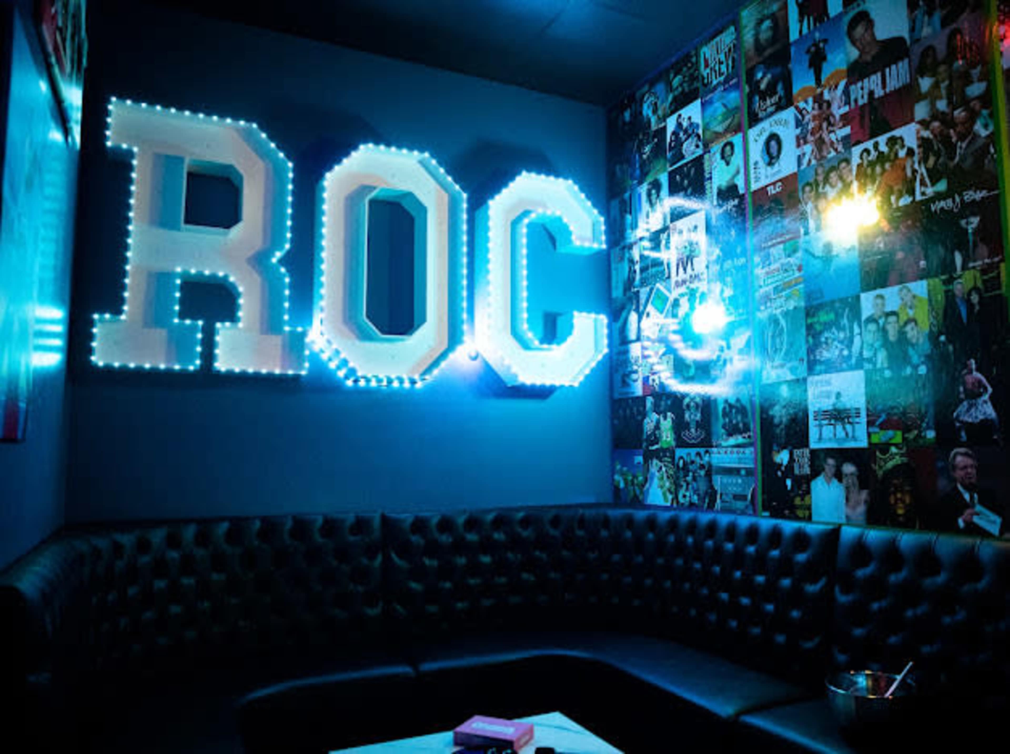 A dimly lit lounge area with a black tufted couch, illuminated by large glowing letters spelling "ROC" and a wall adorned with various music album covers.