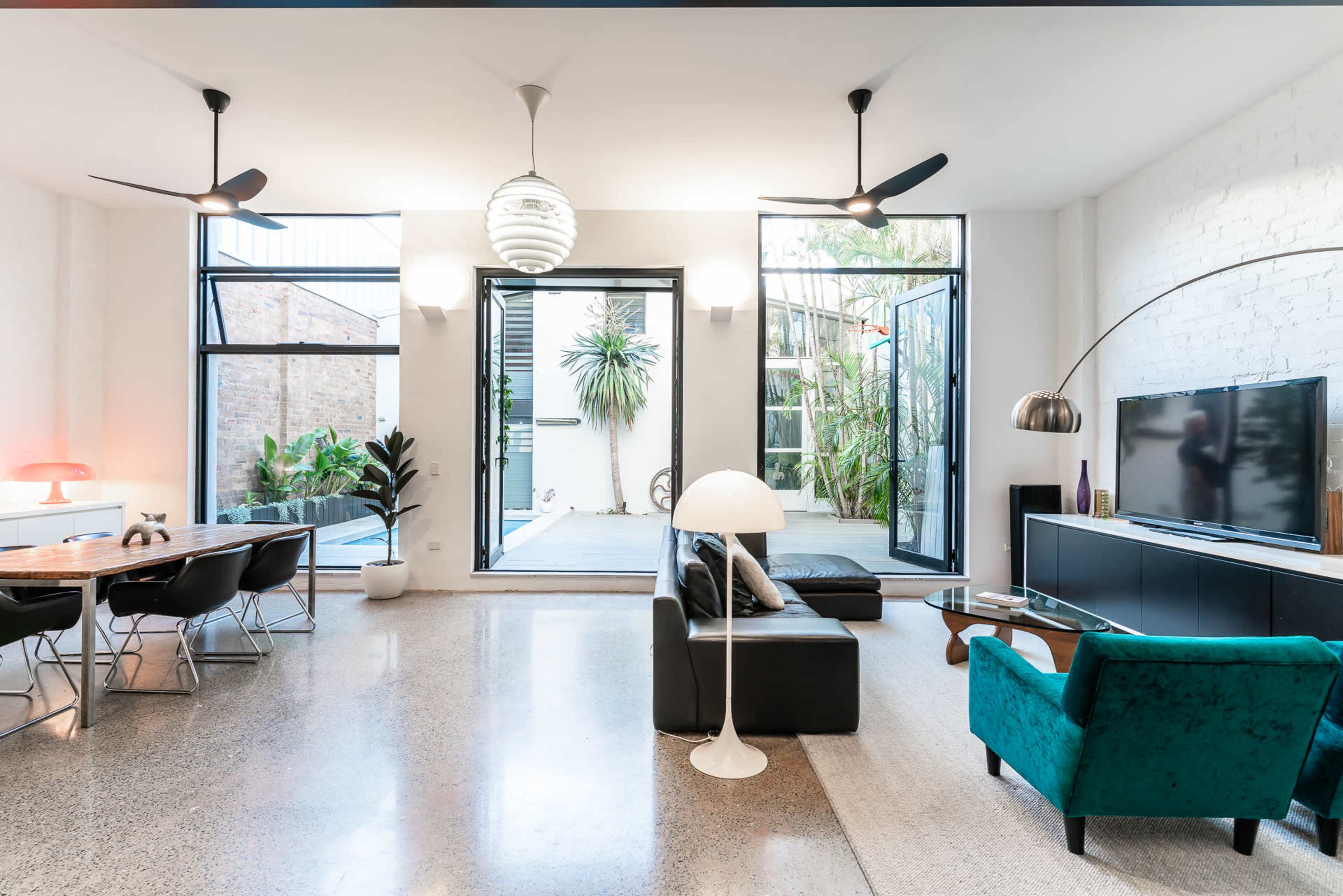 A modern living room featuring a black leather sofa, a green armchair, a large television, and glass doors opening to a patio with plants.