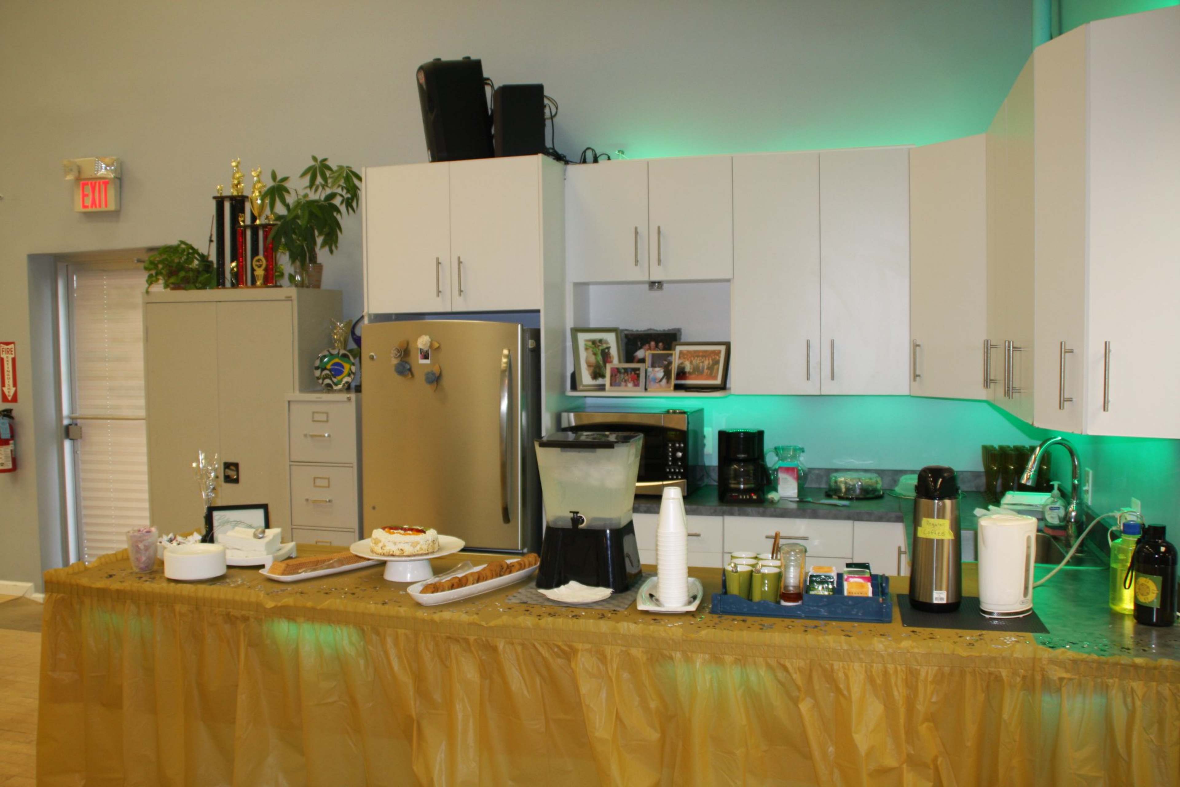 A kitchenette area featuring an assortment of appliances, snacks, and drinks arranged on a counter with a decorative tablecloth and green lighting.