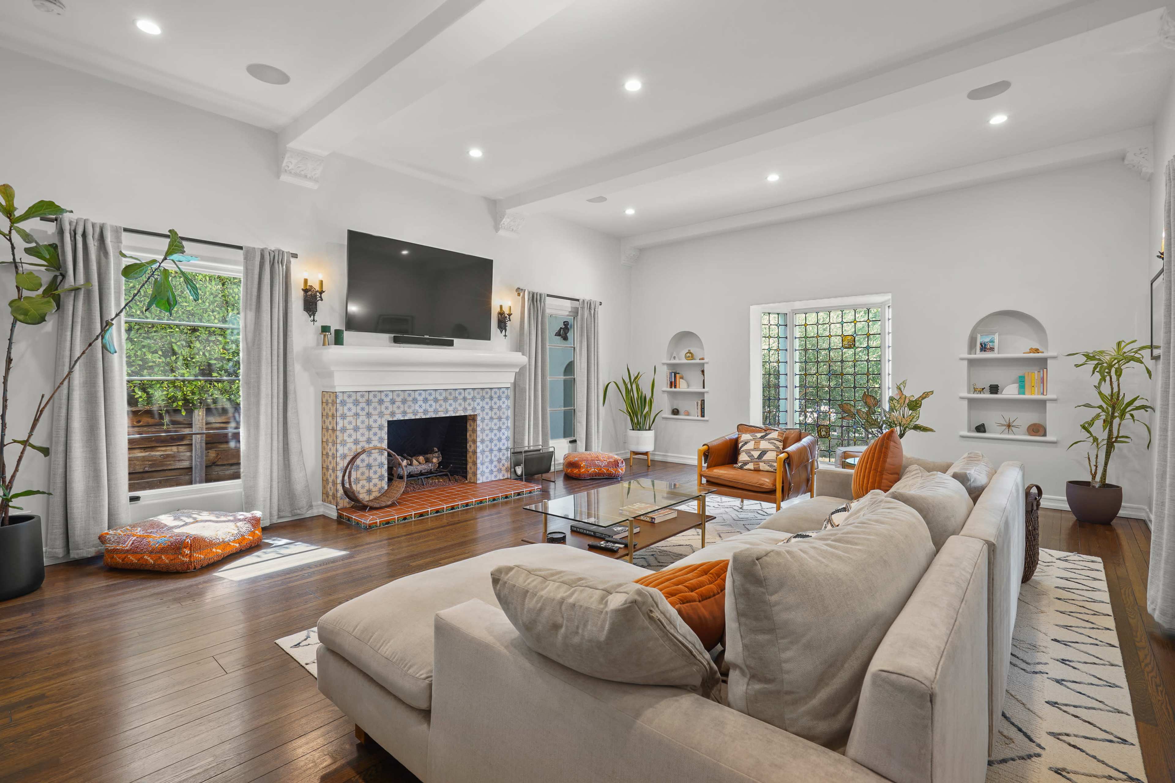 The image features a bright living room with a large sectional sofa, a fireplace, and large windows that open to an outdoor area, accented by decorative shelves and indoor plants.