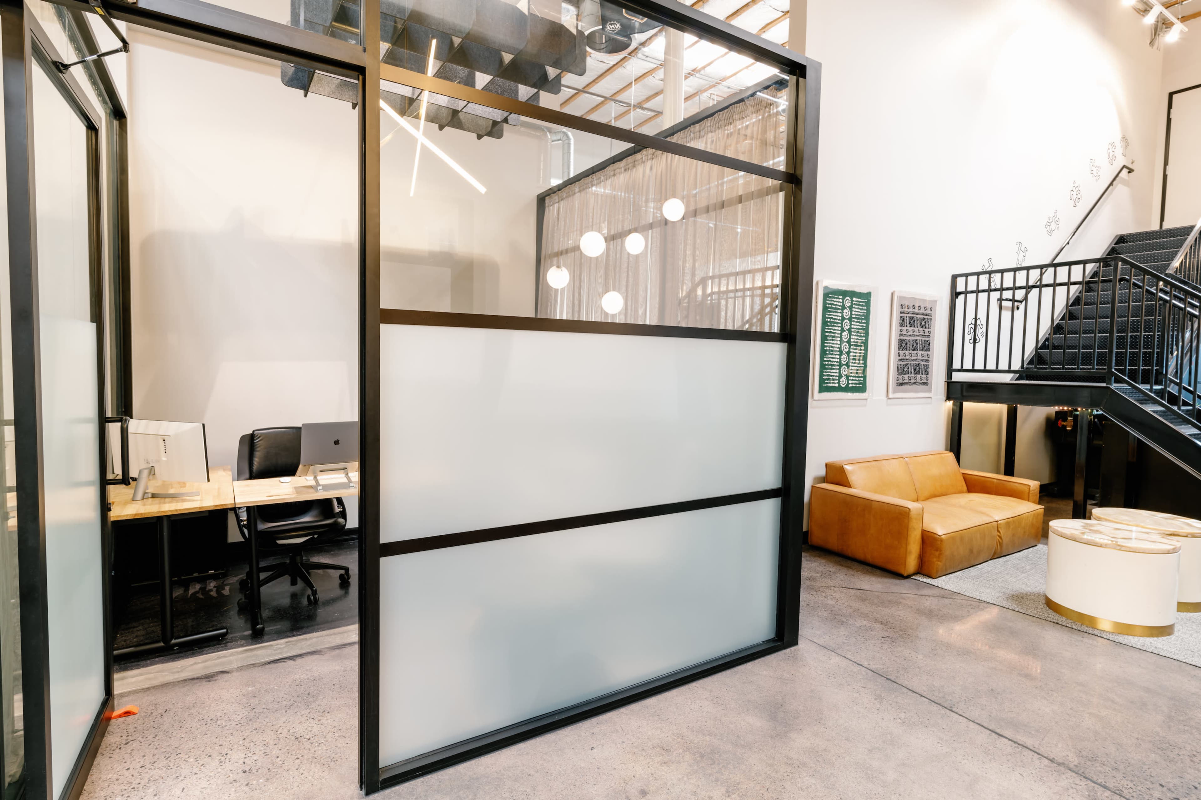 The image shows a modern office space featuring a glass partition, a desk with an ergonomic chair, and a brown sofa near a staircase.