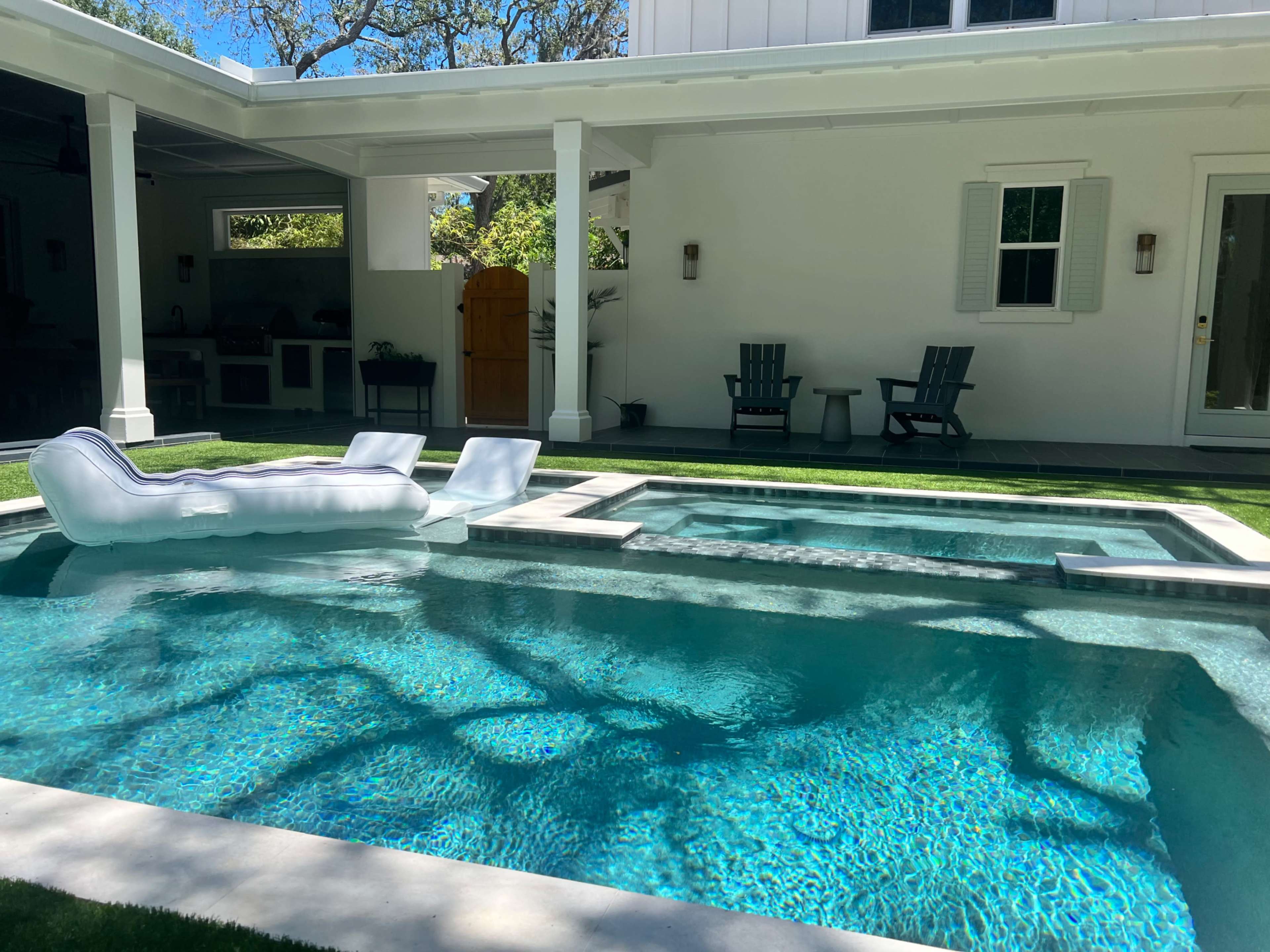 A rectangular swimming pool with a spa area is surrounded by outdoor furniture and the exterior of a house.